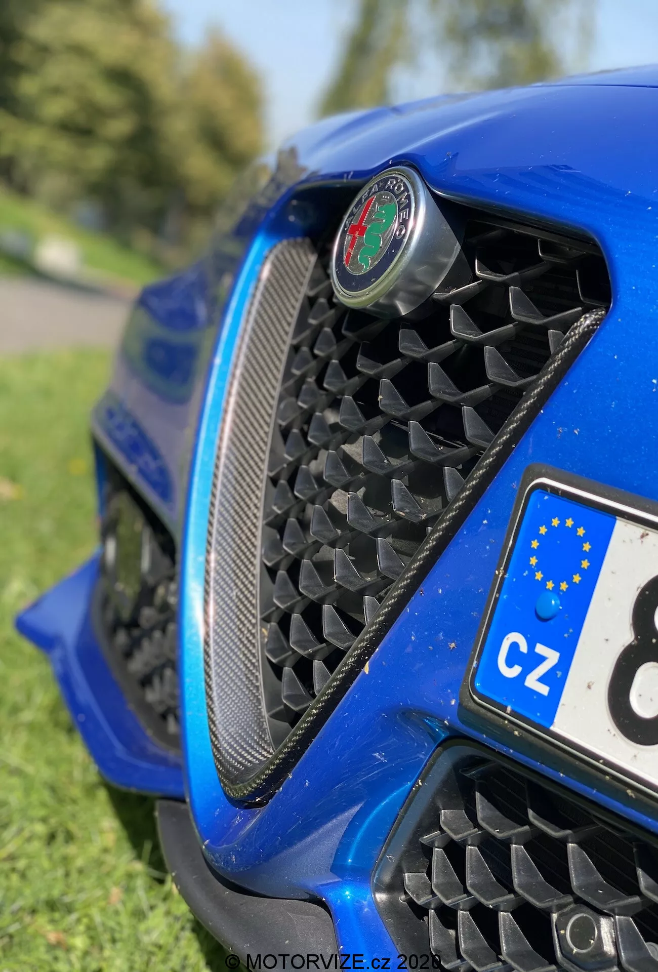 Vista em close-up da parte frontal do Alfa Romeo Giulia 2016 em um ângulo baixo, destacando a grade em forma de colmeia, o emblema central, o para-choque dianteiro aerodinâmico e a carroceria azul metálica brilhante com reflexos. Vista parcial de uma placa de licença tcheca e fundo desfocado semelhante a um parque.