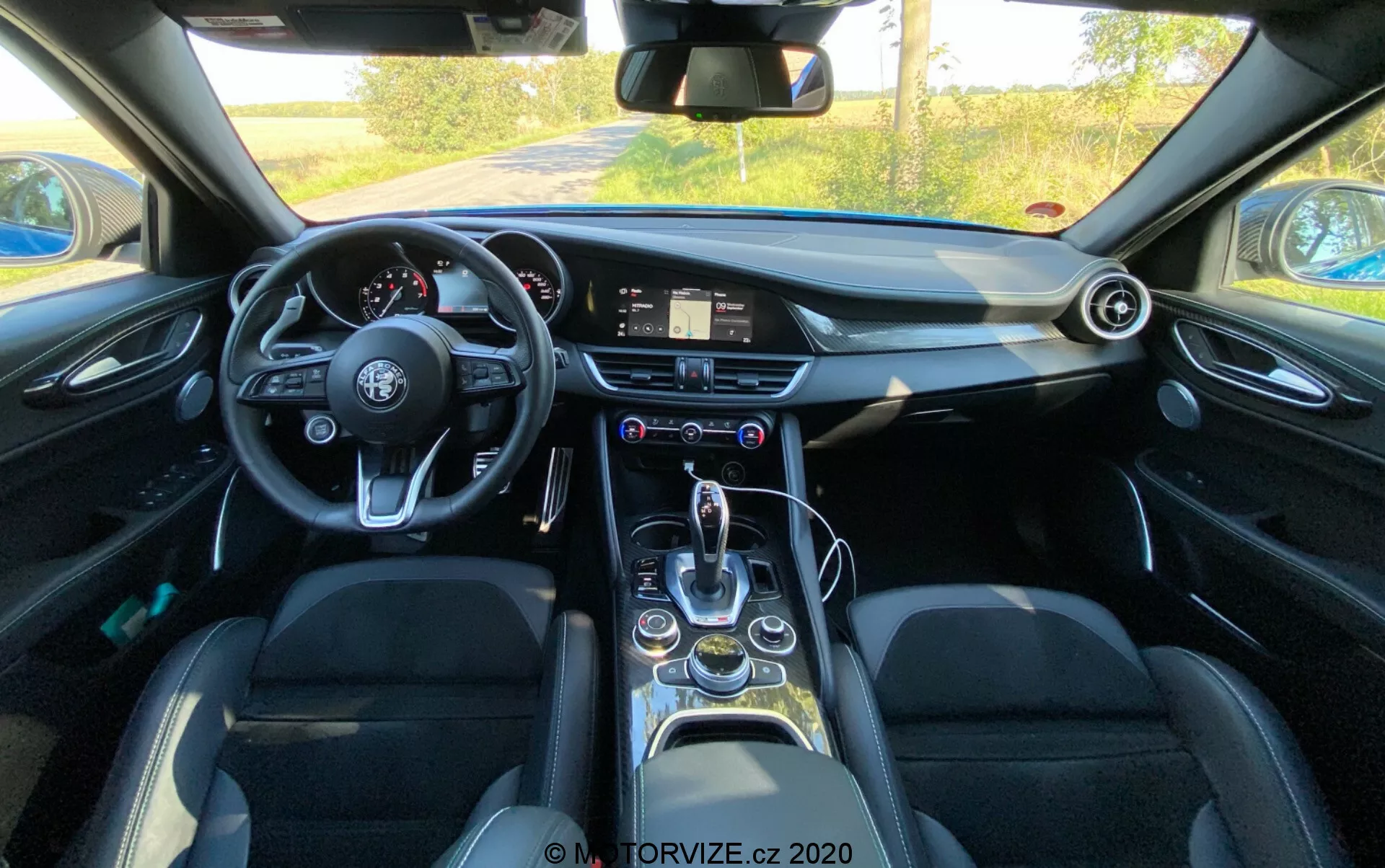 Vista do interior de um Alfa Romeo Giulia 2016 a partir da perspectiva do condutor, mostrando um volante de fundo plano com vários botões de controle, um sistema de infoentretenimento digital no painel de instrumentos, um relógio analógico, ventiladores circulares, uma alavanca de mudanças elegante, seletor de modos de condução, suportes para copos, janelas elétricas, assentos de couro com reforços laterais e características tecnológicas modernas em geral.