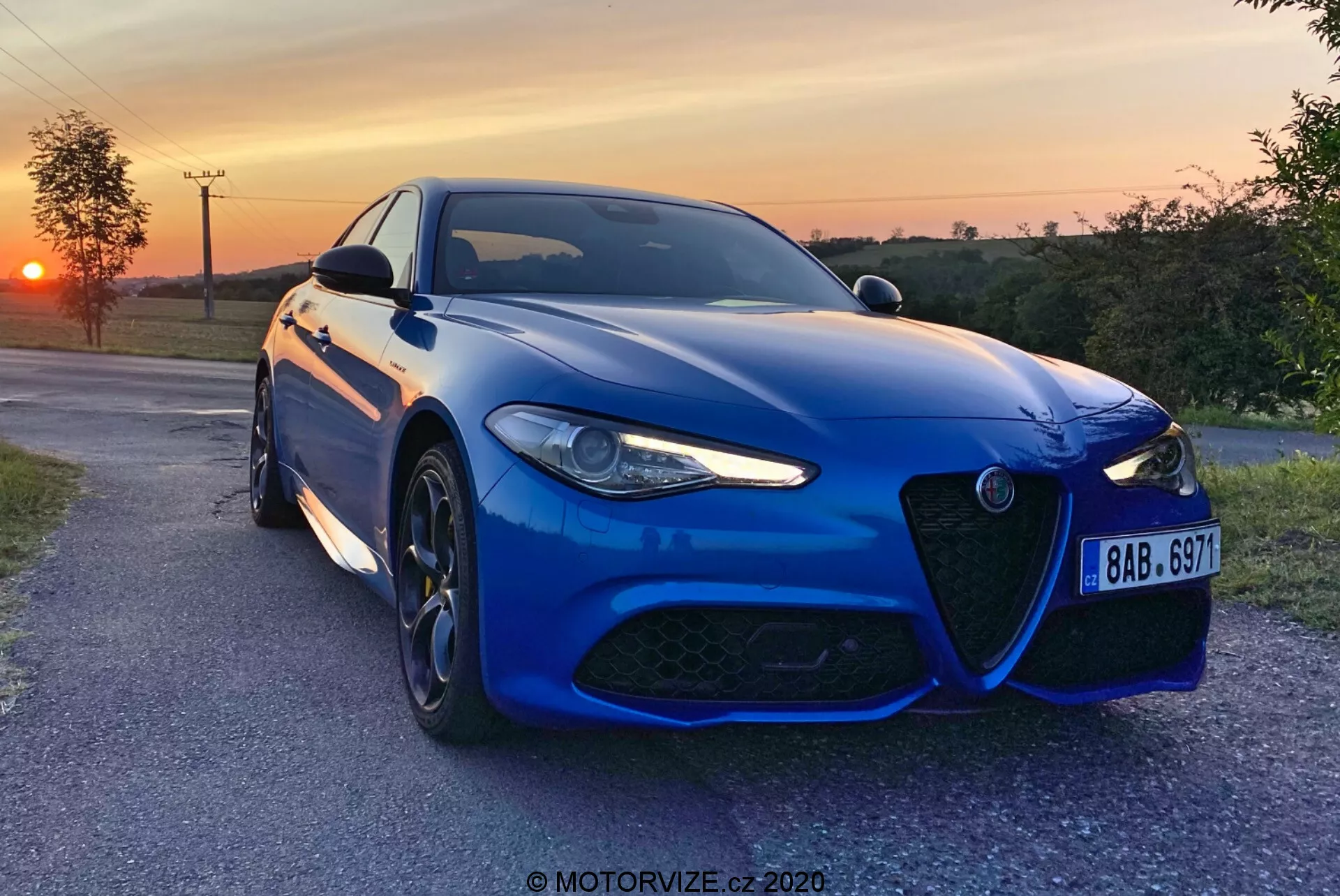 Um sedã esportivo Alfa-Romeo Giulia 2016 azul capturado à noite com um pôr do sol ao fundo, posicionado levemente angular em direção à câmera em uma estrada pavimentada no campo, mostrando sua distintiva grade frontal, design agressivo dos faróis de LED, grandes rodas de liga leve multirraiadas e sistema de freios de alto desempenho. A pintura metálica, o para-choque dianteiro aerodinâmico, as linhas esculpidas do capô e os sensores avançados de assistência ao motorista destacam a estética esportiva e a tecnologia moderna do carro. Uma marca d'água 'MOTORVIZE.cz 2020' está presente.