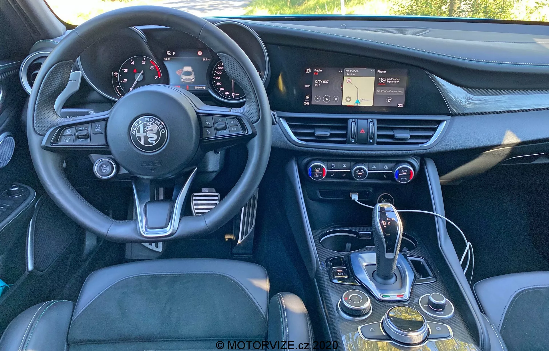 Vista interior de um Alfa Romeo Giulia 2016 de um ângulo ligeiramente desalinhado, de trás para a frente, mostrando o painel de instrumentos, volante, console central e parte do banco do motorista. A imagem destaca o volante de três raios com o emblema da Alfa Romeo, um painel de instrumentos com mostradores analógicos e displays digitais, um painel com um sistema de infoentretenimento com tela sensível ao toque, saídas de ar e controles de clima e multimídia. O console central inclui um seletor de transmissão automática, um disco giratório, botões para navegação de infoentretenimento e configurações do veículo, e um possível seletor de modos. O design enfatiza ergonomia e acessibilidade, com materiais como couro, metal escovado e plástico de alta qualidade para uma sensação de luxo.