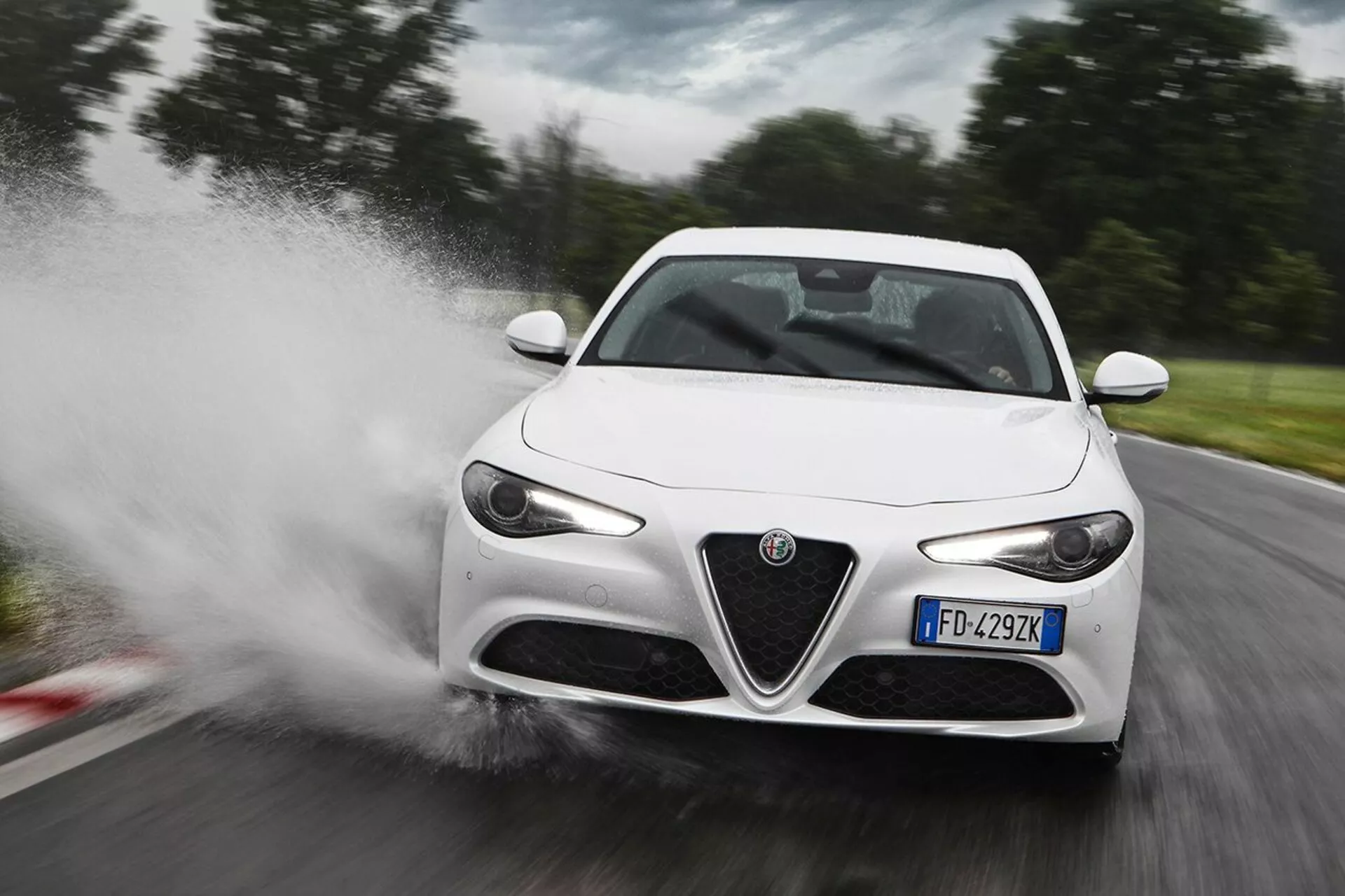 Carro esportivo Alfa Romeo Giulia branco de 2016 acelerando através da água, causando respingos, em vista frontal de três quartos destacando seus faróis LED, grade frontal distinta, espelhos laterais elegantes e linhas aerodinâmicas
