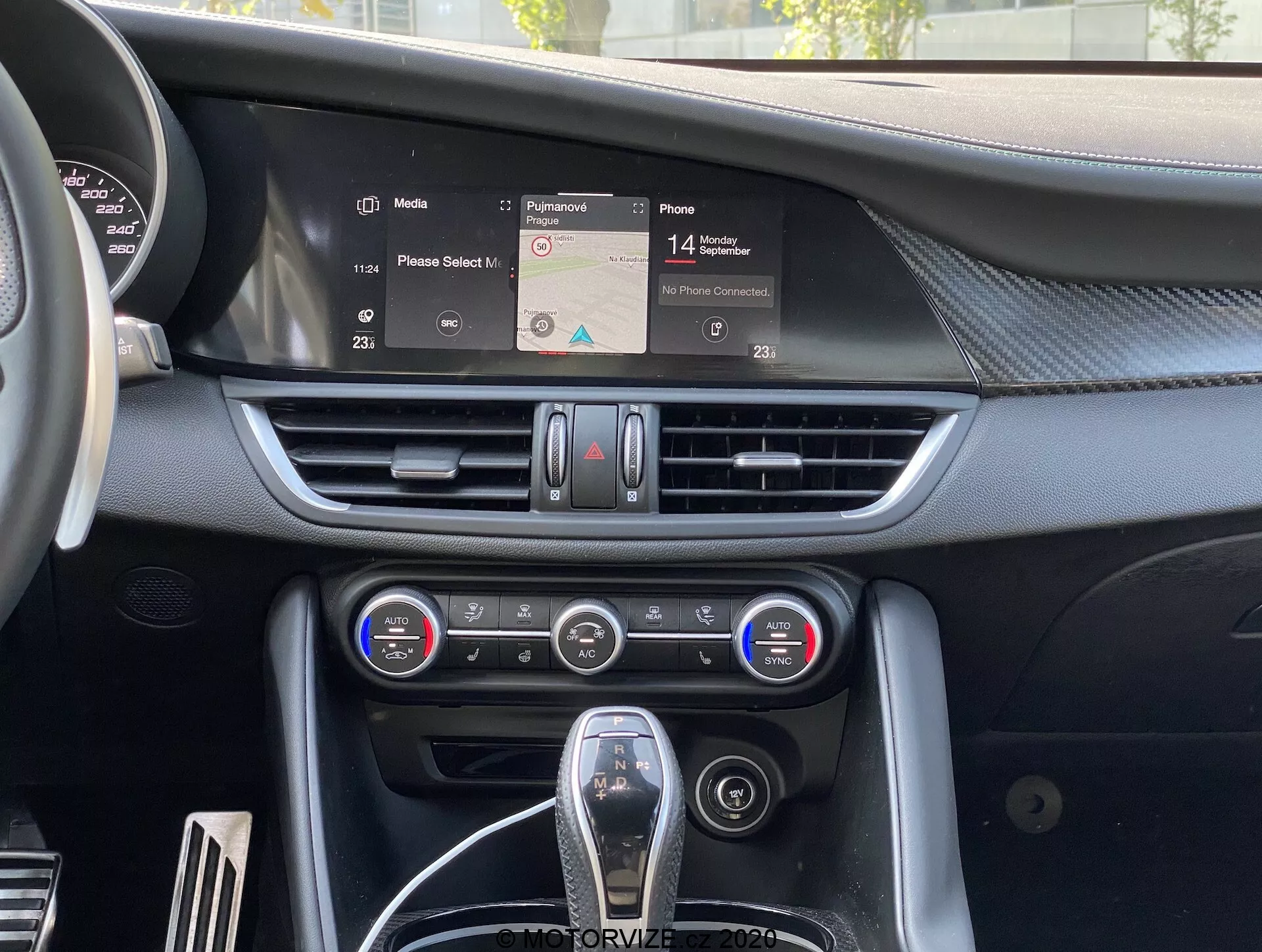 Vista interna do Alfa-Romeo Giulia 2016 focando no painel central com uma tela de infoentretenimento exibindo um mapa de navegação, saídas de ar horizontais, painel de controle de climatização de duas zonas com botões e dials, e uma alavanca de câmbio parcialmente visível com acabamento em couro e detalhes cromados, representando a ergonomia e tecnologia moderna do interior do carro.