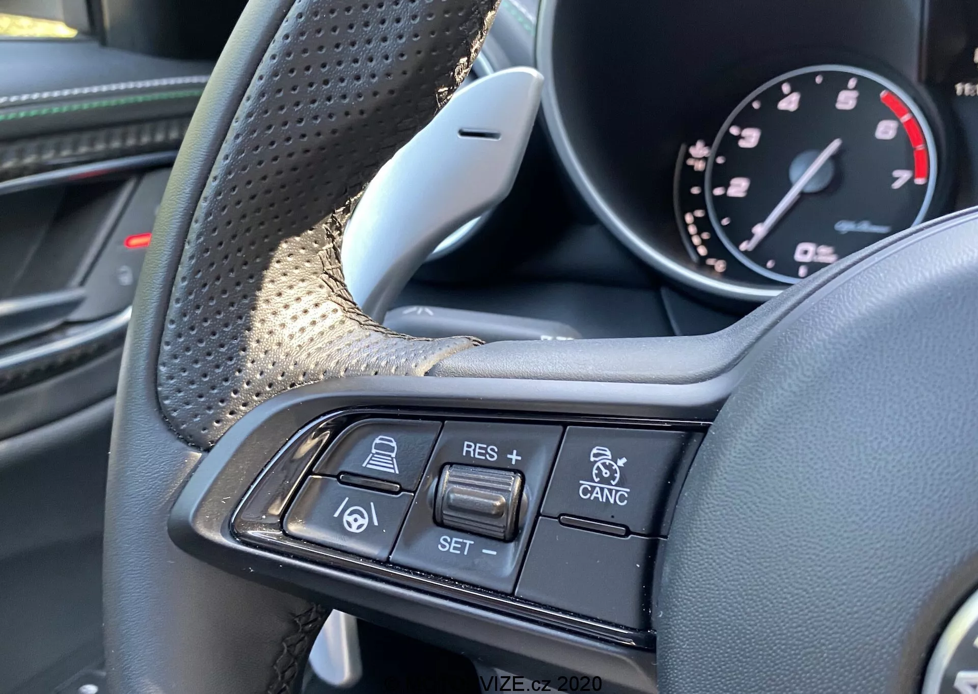 Vista aproximada do interior do Alfa-Romeo Giulia 2016, destacando o volante e seus controles multifuncionais a partir da perspectiva do motorista. As áreas texturizadas de aderência e materiais luxuosos como couro e possivelmente fibra de carbono são visíveis no lado esquerdo. O lado direito apresenta botões do controle de cruzeiro e tecnologia de cruzeiro adaptativo, incluindo + RES, - SET, CANCEL, um ícone de carro seguindo carro e alertas de colisão de veículos. A imagem concentra-se exclusivamente nesses controles, destacando o design premium e recursos avançados de assistência ao motorista.