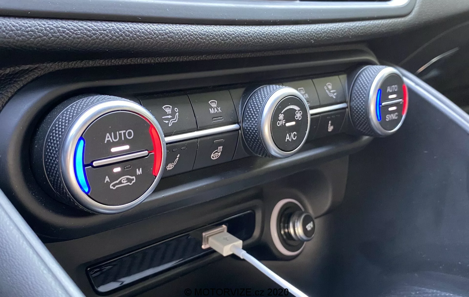 Close-up do sistema de controle climático no painel de instrumentos do Alfa Romeo Giulia 2016, apresentando três botões circulares. O botão esquerdo indica configurações de temperatura com faixas azuis e vermelhas e uma configuração 'AUTO', parcialmente iluminado em vermelho. O botão do meio mostra configurações de velocidade do ventilador, destacado em uma velocidade mais baixa. O botão direito exibe vários modos de distribuição de ar com botões 'AC' e 'SYNC'. Abaixo dos botões há uma tomada de corrente de 12 volts e uma porta USB. O painel tem um acabamento preto com um destaque prateado ao redor dos controles climáticos, capturado de um ângulo elevado.