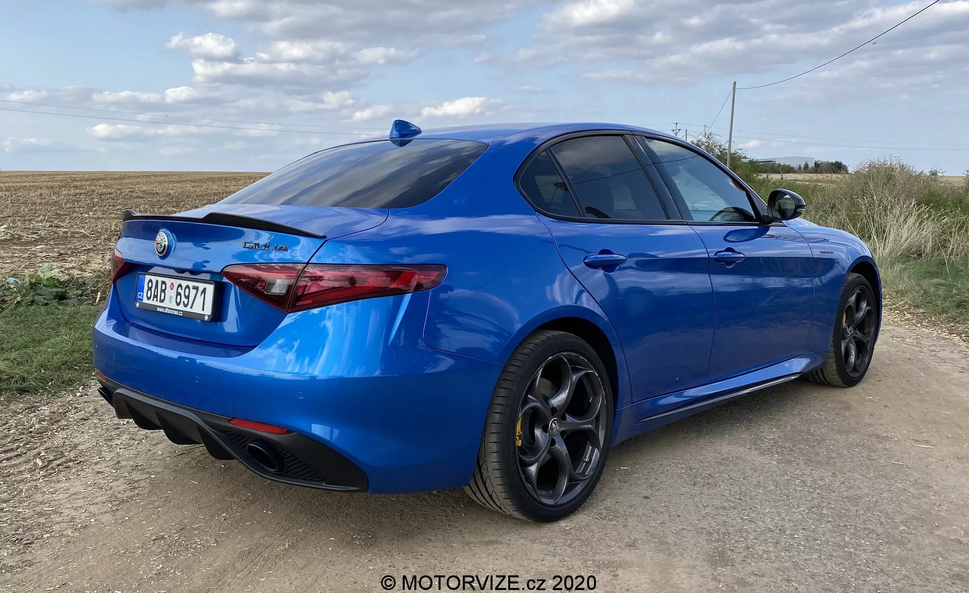 Vista traseira de três quartos de um sedã esportivo azul brilhante em uma estrada não pavimentada com campo aberto e céu parcialmente nublado ao fundo. Fotografia de baixo ângulo destacando a postura do carro, para-choque traseiro esculpido com escapes duplos integrados, conjuntos de luzes traseiras finas e orientadas horizontalmente, spoiler na tampa do porta-malas e rodas de liga leve pretas multifinos com pneus de alto desempenho de perfil baixo. O perfil lateral mostra uma silhueta elegante com linha de teto inclinada e janelas traseiras ligeiramente escurecidas. A traseira do carro apresenta um emblema e símbolo da marca indicando o modelo ou nível de acabamento. O exterior do carro parece estar em excelente estado, e a luz do dia proporciona boa visibilidade do design e da pintura do carro.