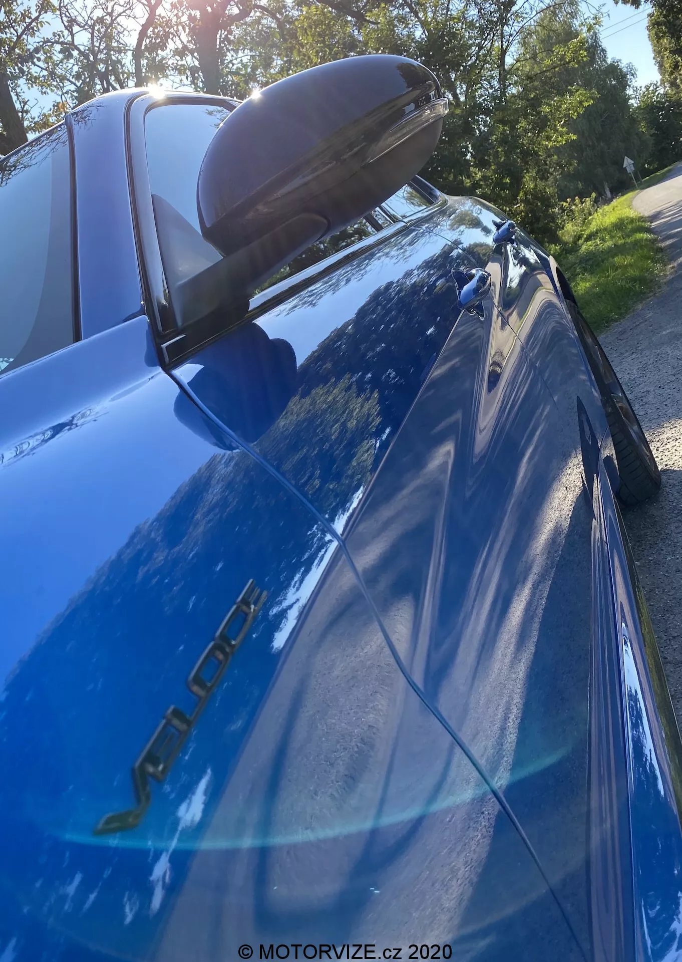 Imagem aproximada de um Alfa Romeo Giulia azul (modelo 2016) fotografada de um ângulo baixo e para cima focando na seção traseira. O emblema do modelo no lado do motorista, a pintura azul refletiva, a luz traseira esquerda de LED, o spoiler traseiro preto, a janela traseira inclinada com a terceira luz de freio, o reflexo de árvores e céu brilhante e o espelho lateral com sinalizador integrado são visíveis.