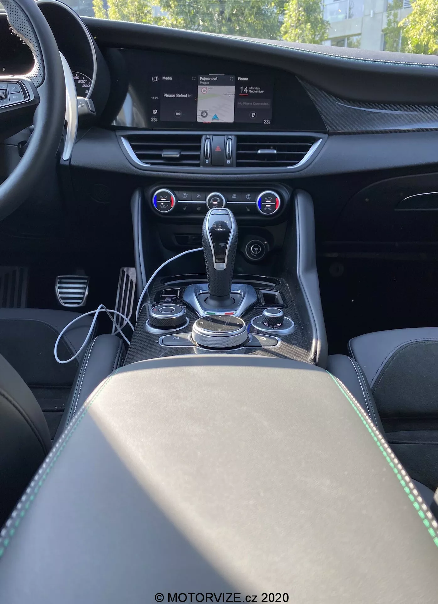 Imagem do interior do Alfa Romeo Giulia 2016 vista de trás da consola central, mostrando a pilha central, o túnel de transmissão e parte do assento do passageiro. A imagem oferece uma perspectiva frontal do ponto de vista de um passageiro traseiro, destacando uma alavanca de câmbio automática moderna rodeada por botões e controles metálicos para o sistema de infotainment e modos de condução. Acima da área da alavanca de câmbio está o sistema de controle climático automático de duas zonas com um display digital mostrando as configurações de temperatura e botões para controlar a distribuição de ar e a velocidade do ventilador. Uma grande tela de infotainment no topo da pilha central exibe o mapa do sistema de navegação. O painel apresenta detalhes de costura e acabamento em aspecto metálico, sugerindo materiais premium. O apoio de braço e o compartimento de armazenamento com dois porta-copos são visíveis atrás da alavanca de câmbio, enriquecendo o design luxuoso e focado em tecnologia da cabine.