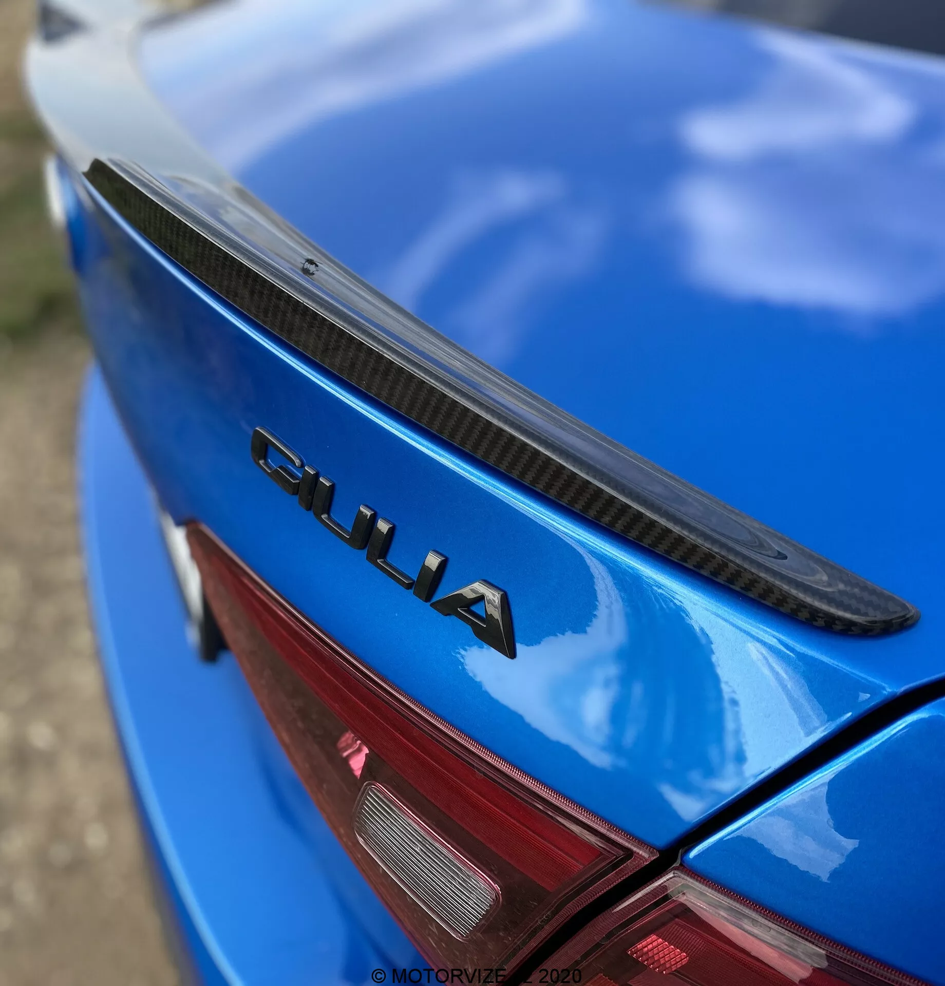Vista em close-up e angulada da seção traseira de um Alfa Romeo Giulia 2016 azul. A fotografia mostra a tampa do porta-malas com um spoiler preto, pintura azul brilhante e um emblema cromado 'GIULIA' destacado. Uma parte da luz traseira LED vermelha também é visível. O emblema 'GIULIA' está nitidamente focado, enquanto o spoiler, a superfície do carro e a luz traseira se tornam gradualmente desfocados, destacando os elementos de marca e design do carro.