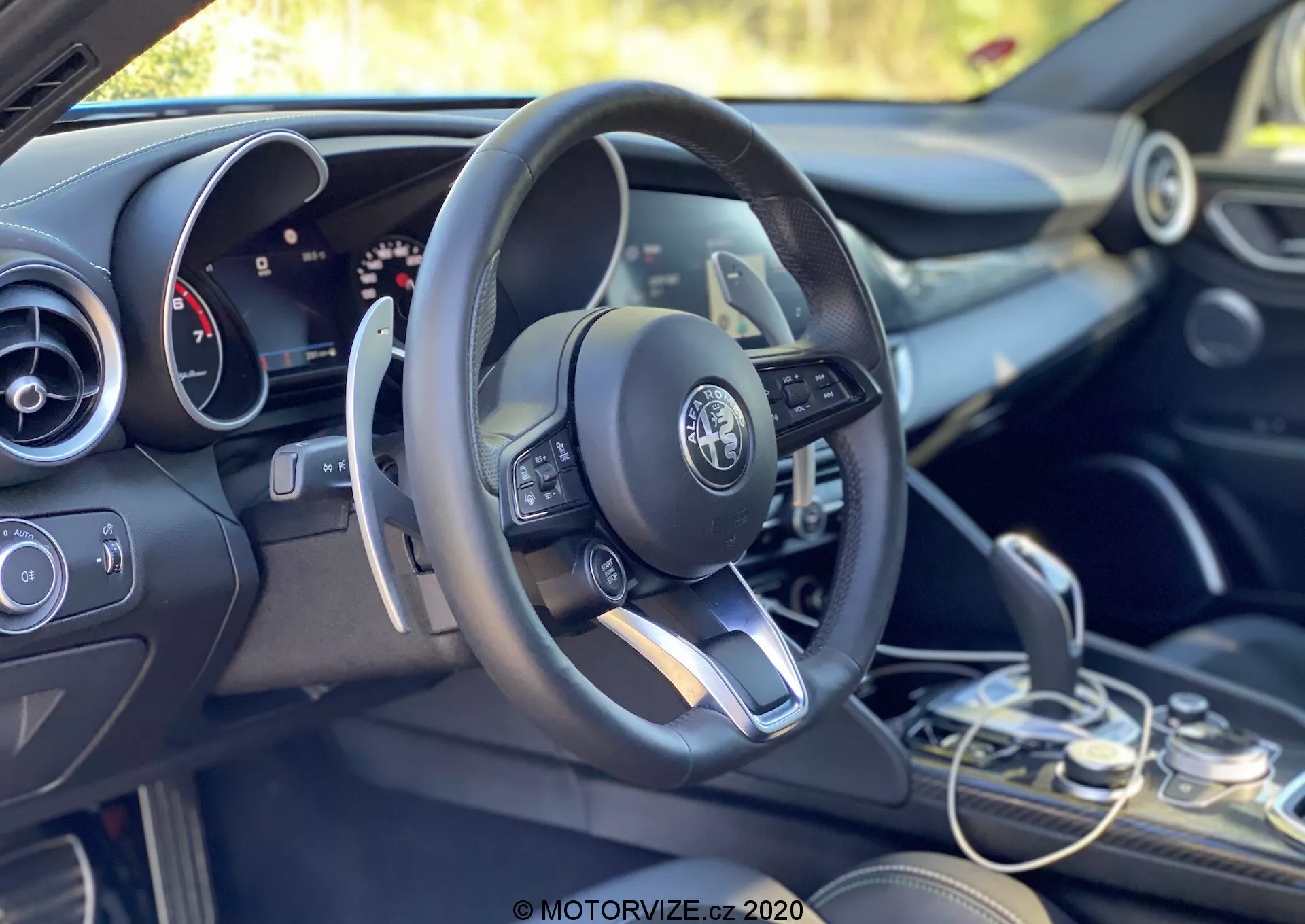 Vista interior de um Alfa Romeo Giulia 2016 focada na área do condutor e no painel a partir de uma perspectiva inclinada para trás, do lado esquerdo. A configuração de condução à esquerda apresenta um volante multifuncional moderno com controles montados, incluindo botões para ajuste de volume, acesso a menus e, possivelmente, controle de cruzeiro. O logotipo da Alfa Romeo é claramente visível no centro do volante. Atrás do volante, parcialmente visível, está um painel de instrumentos misto com exibições analógicas e digitais, indicando um sistema de informações do condutor configurável. À direita do volante, o topo da tela do sistema de infotainment é visível, ladeada abaixo pelos controles do sistema de clima e interruptores funcionais. O seletor de marchas está localizado no canto inferior direito, mostrando um design elegante para transmissão automática. Os acabamentos internos combinam tons escuros com acentos metálicos, refletindo o design interno premium do veículo. A cabine está banhada por luz natural, com um fundo verde sugerindo um ambiente exterior ou vegetativo.