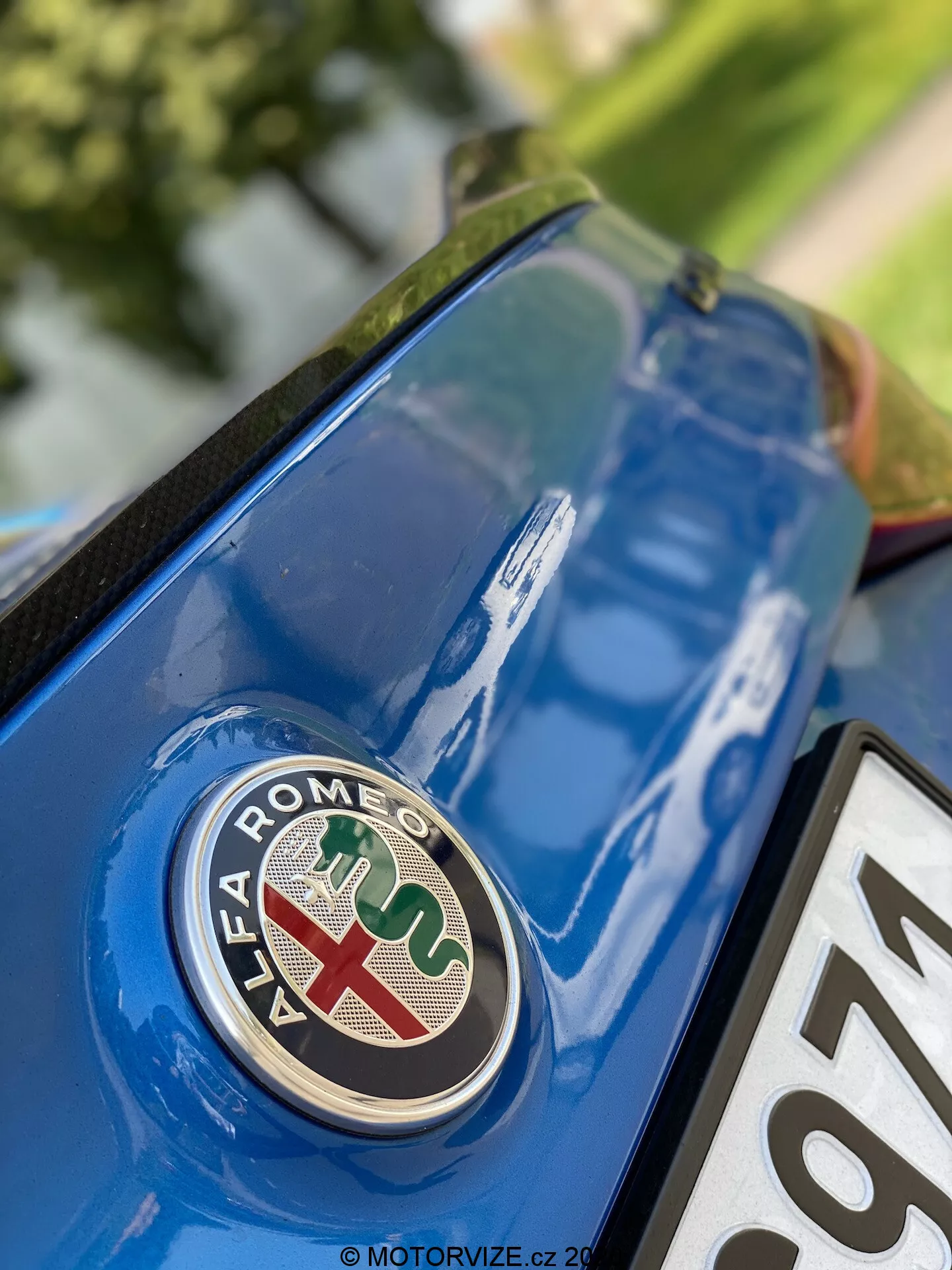 Vista aproximada da seção traseira do Alfa Romeo Giulia 2016 com foco no emblema da marca a partir de um ângulo baixo. O distintivo, com uma cruz vermelha sobre fundo branco e uma cobra verde sobre fundo azul, está em nítido foco na tampa do porta-malas azul metálica, com detalhes e reflexos da superfície circundante.