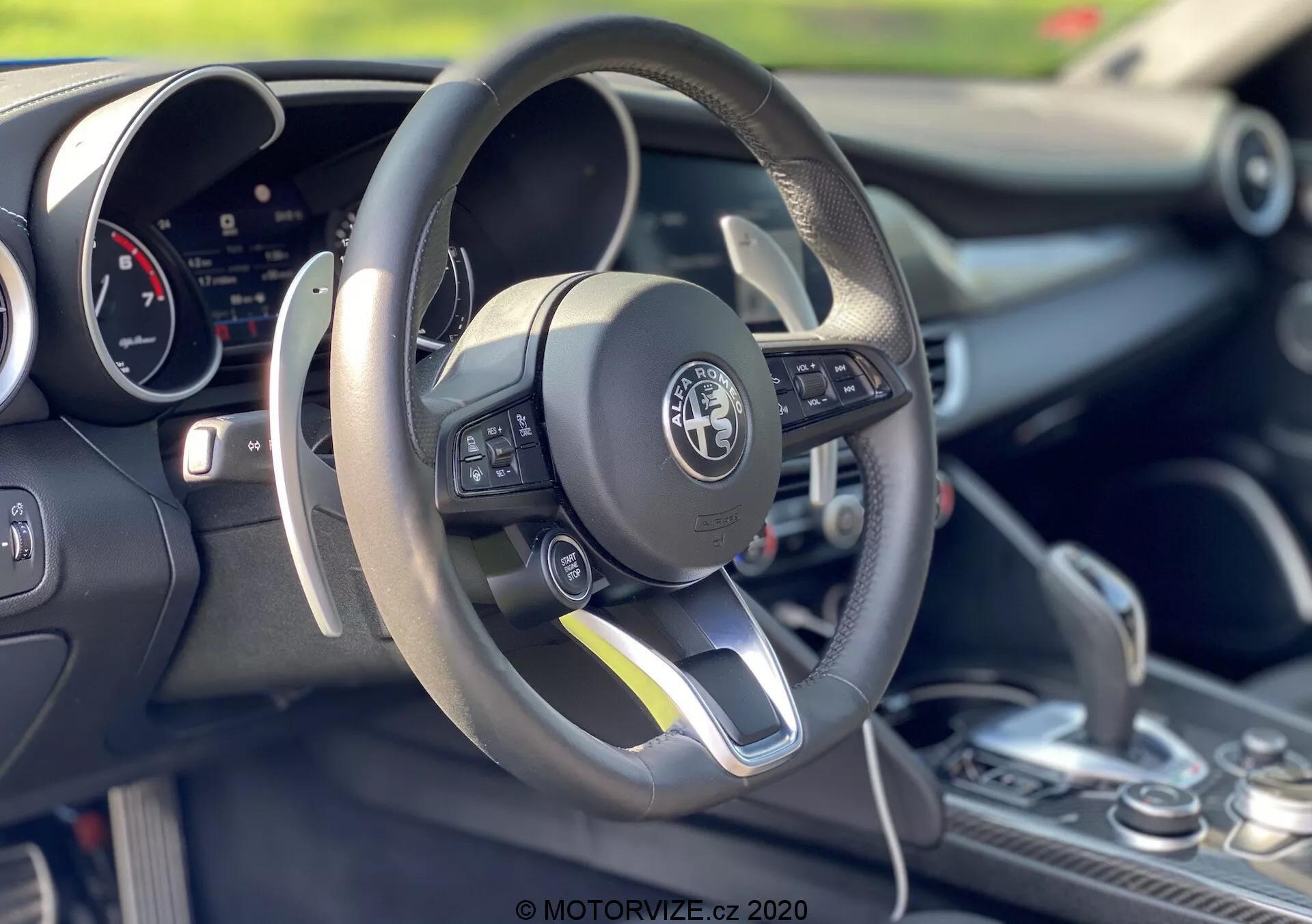Imagem interior de um Alfa-Romeo Giulia 2016 focando na área do motorista e no volante. O volante de fundo plano revestido em couro apresenta controles multifuncionais no lado esquerdo. Atrás do volante, está visível um painel de instrumentos digital. O console central mostra uma alavanca de câmbio com uma cobertura de couro, sugerindo possíveis capacidades de troca manual. A imagem enfatiza o design esportivo do carro, tecnologia avançada e artesanato detalhado com uma profundidade de campo rasa destacando os elementos em primeiro plano.