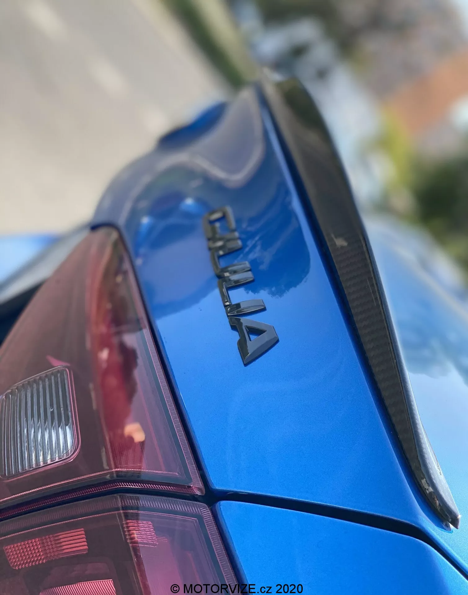 Imagem em close-up da seção traseira de um carro azul ŠKODA, vista de um ângulo para cima. O foco está no logotipo de cromo 'ŠKODA' na tampa do porta-malas, um spoiler fino e a pintura brilhante e bem conservada. O para-brisa traseiro possui linhas de desembaçador e parte do conjunto de luz traseira moderna é visível. A baixa profundidade de campo enfatiza a marca, com a parte frontal e o fundo desfocados. Com marca d'água 'MOTORVIZE.cz 2020', indicando a data de publicação ou captura.