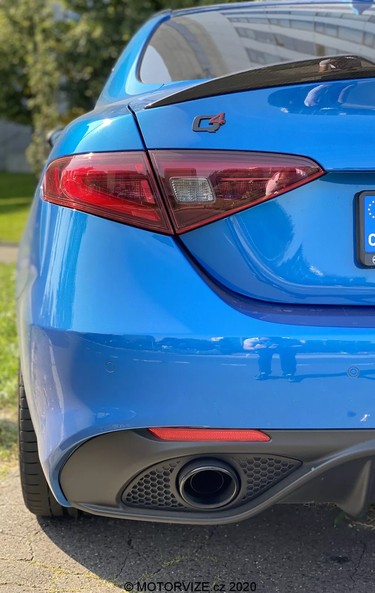 Close-up da seção traseira de um carro azul mostrando o lado direito a partir de um ângulo baixo de três quartos. O enquadramento captura o emblema proeminente 'C4' na tampa do porta-malas, uma elegante lanterna traseira de LED, pintura azul vibrante, um único tubo de escape cromado e o difusor integrado no para-choque traseiro. A pequena profundidade de campo destaca o emblema e a lanterna traseira, enquanto a luz natural do sol se reflete nas curvas suaves e esportivas da carroceria do carro.