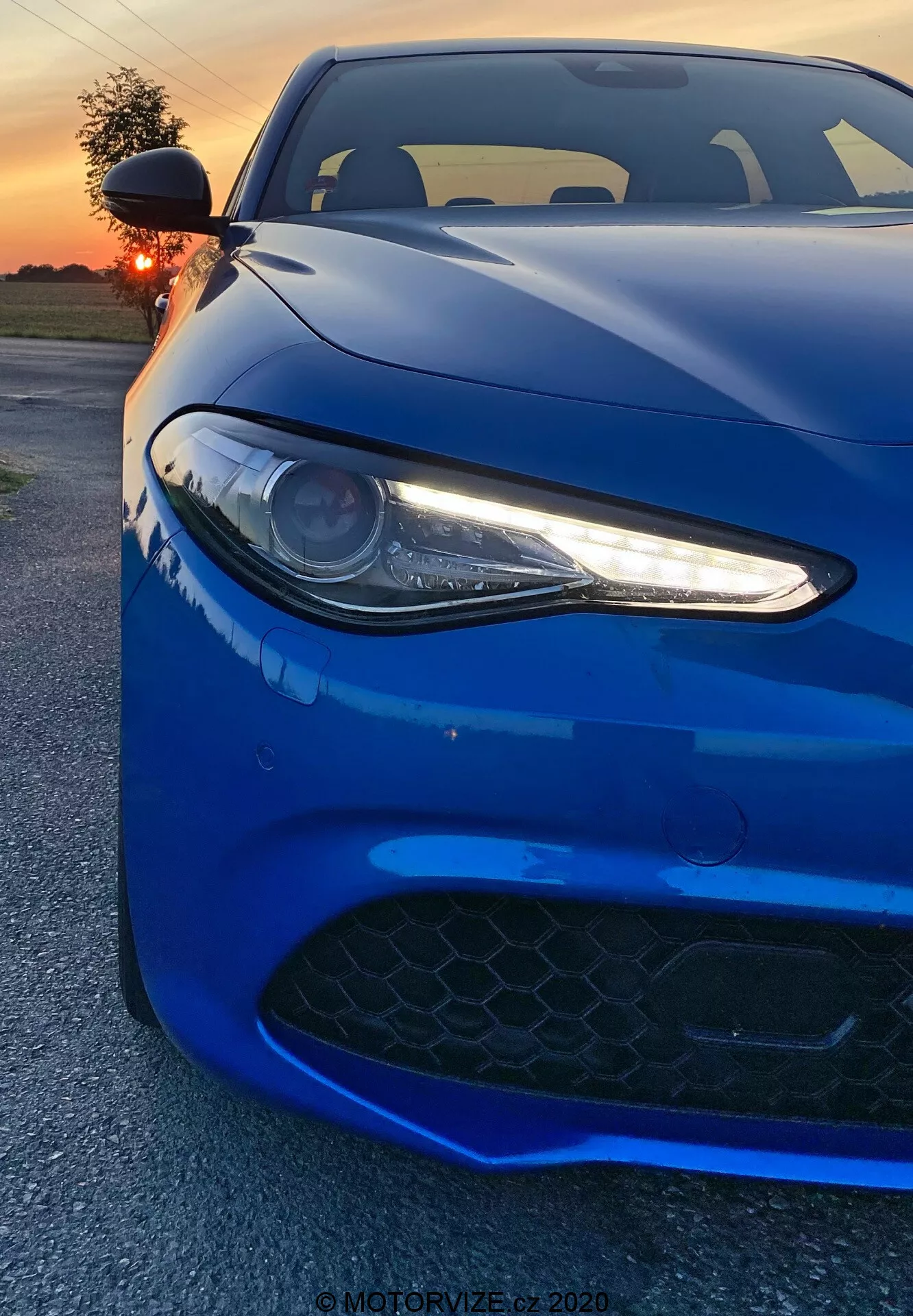 A imagem mostra um carro esportivo Alfa Romeo Giulia 2016 capturado de um ângulo frontal baixo. O vibrante veículo azul profundo preenche o quadro, enfatizando seu farol esquerdo com luzes diurnas de LED, parte do capô e a grade de entrada de ar no para-choque dianteiro. O farol do tipo projetor, com seus elegantes acentos LED brancos, confere uma aparência agressiva e moderna. A grade preta com padrão hexagonal sugere capacidades de alto desempenho. A tomada captura as linhas e contornos aerodinâmicos do carro, com reflexos do pôr-do-sol realçando seu acabamento brilhante. Ao fundo, vê-se um céu laranja iluminado parcialmente oculto pelo sol. O ângulo próximo do fotógrafo destaca os detalhes do design, evocando uma sensação de poder e elegância.
