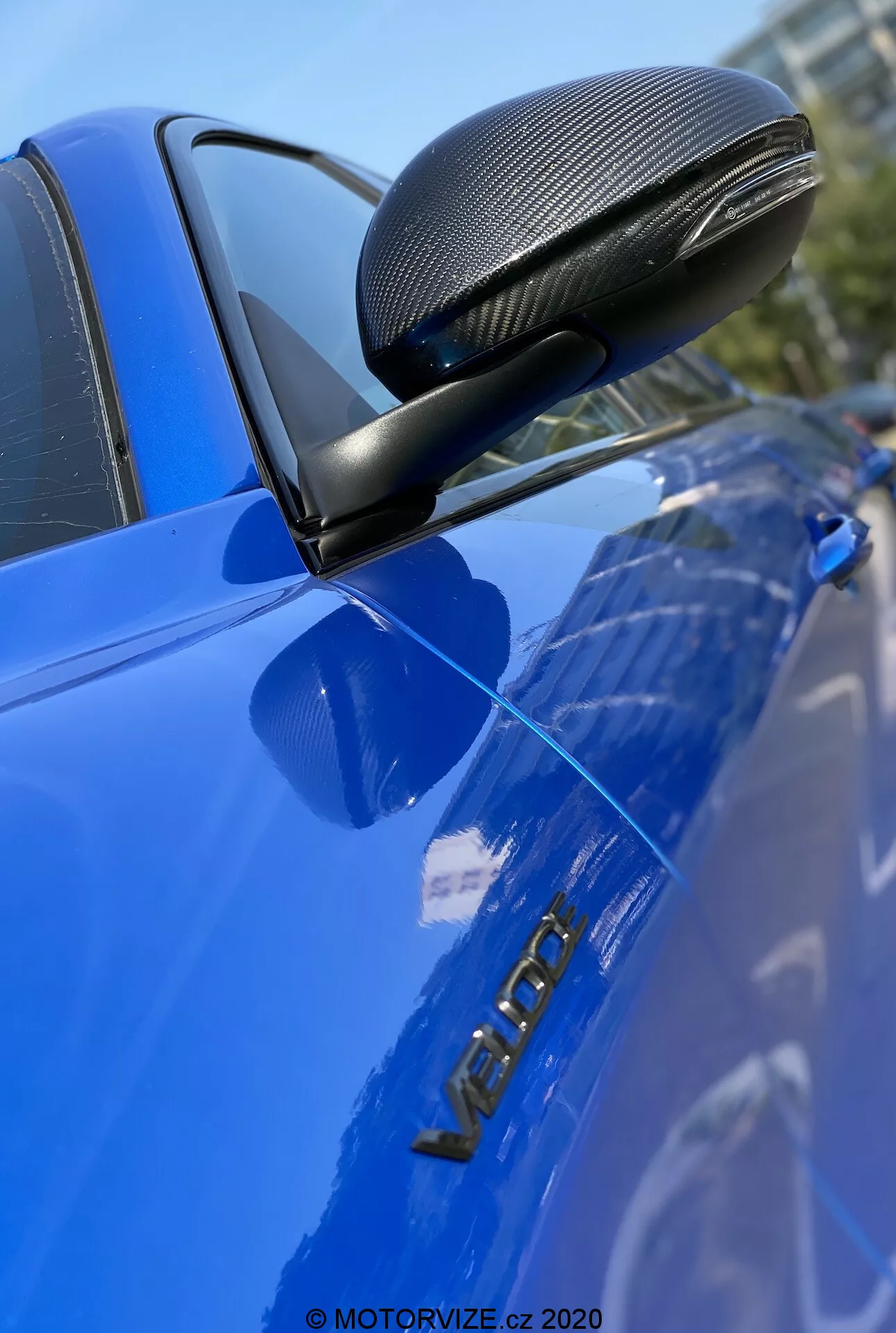 Imagem em close-up de um carro Alfa Romeo Giulia 2016 azul, enfatizando o retrovisor lateral com acabamento em fibra de carbono em uma carcaça aerodinâmica, montado em um suporte preto. A foto foca nitidamente no espelho, na porta do motorista e na insígnia metálica 'VERON', destacando esses detalhes com pouca profundidade de campo e uma superfície brilhante e bem conservada.