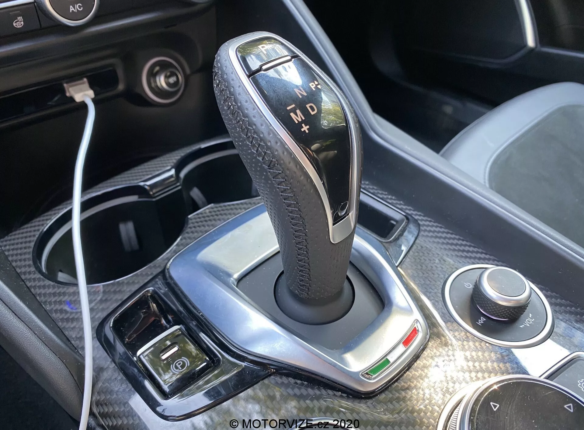 Fechamento do alavanca de câmbio da transmissão automática do Alfa-Romeo Giulia 2016, destacando uma moldura prateada polida, empunhadura preta texturizada de couro e indicadores iluminados para 'P', 'R', 'N', 'D' e 'M'. A luz do dia brilhante realça o design, com um dial rotativo e conectividade USB visíveis nas proximidades. Acabamento cromado, indicador verde e botões adicionais para freio de estacionamento eletrônico e controle de clima são visíveis, oferecendo uma visão do luxo e da tecnologia avançada do interior do carro.