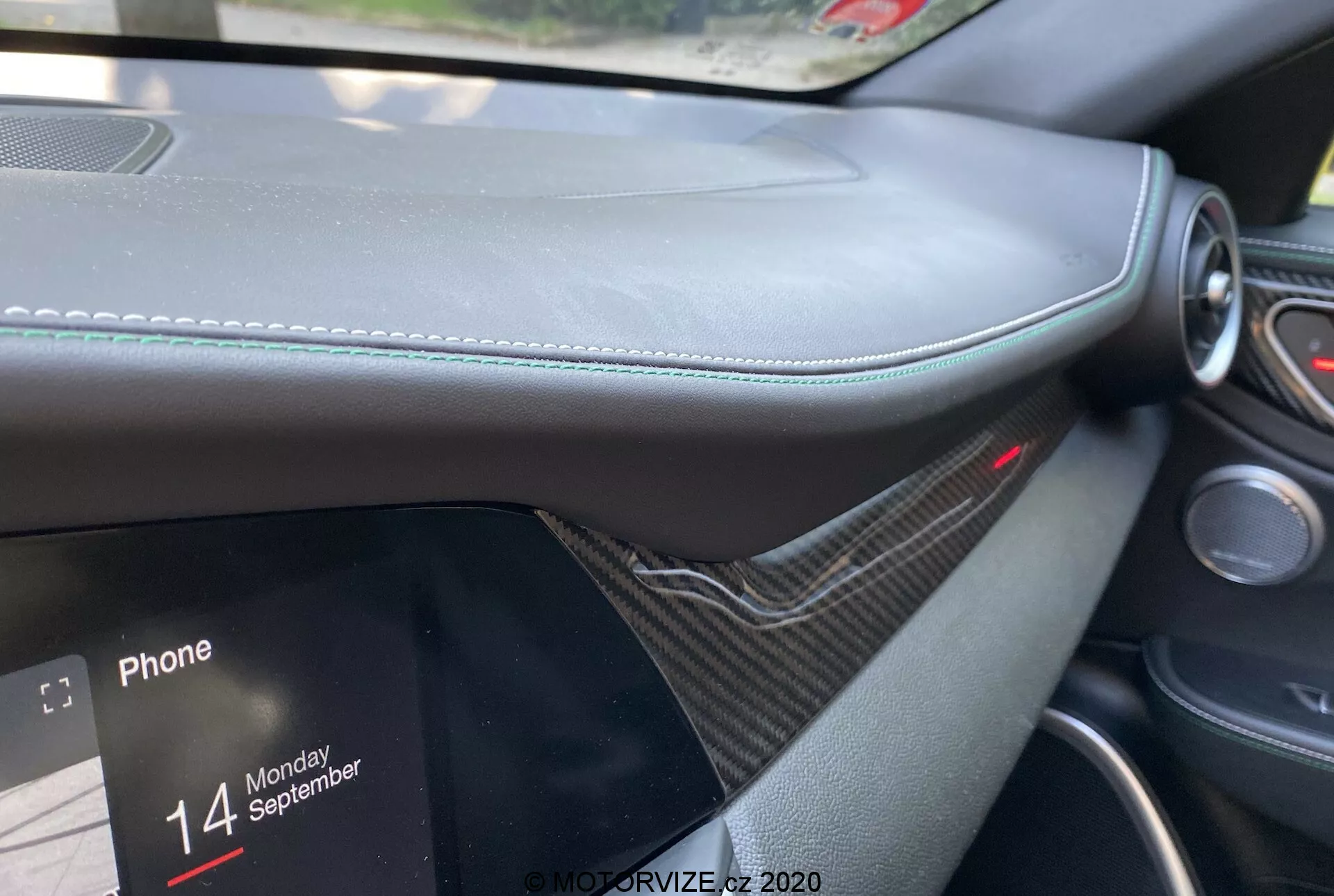 Close-up do interior do Alfa Romeo Giulia 2016 a partir da perspectiva do passageiro, focando no painel com material semelhante a couro e costuras visíveis, acabamento semelhante a fibra de carbono, tela de infoentretenimento parcialmente visível exibindo opções de conectividade telefônica com o texto 'Phone' e 'Monday 14 September'. Luz vermelha acima do sistema de infoentretenimento indicando um sistema de segurança ativo, enfatizando luxo e design moderno.