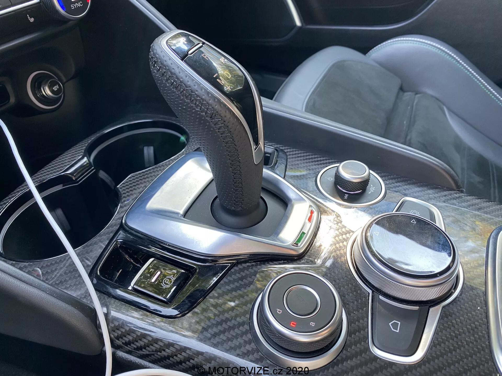 Vista de perto da consola central e da área da alavanca de mudanças do Alfa Romeo Giulia 2016 com uma alavanca de mudanças automática revestida a couro com detalhes cromados, rodeada por acabamentos em fibra de carbono. A consola inclui um botão de controlo tipo iDrive, botão de controlo de volume, botões de dinâmica de condução, porta USB com cabo conectado e um painel com botões de emergência e outros, enfatizando o design ergonómico e tecnológico do interior do carro.