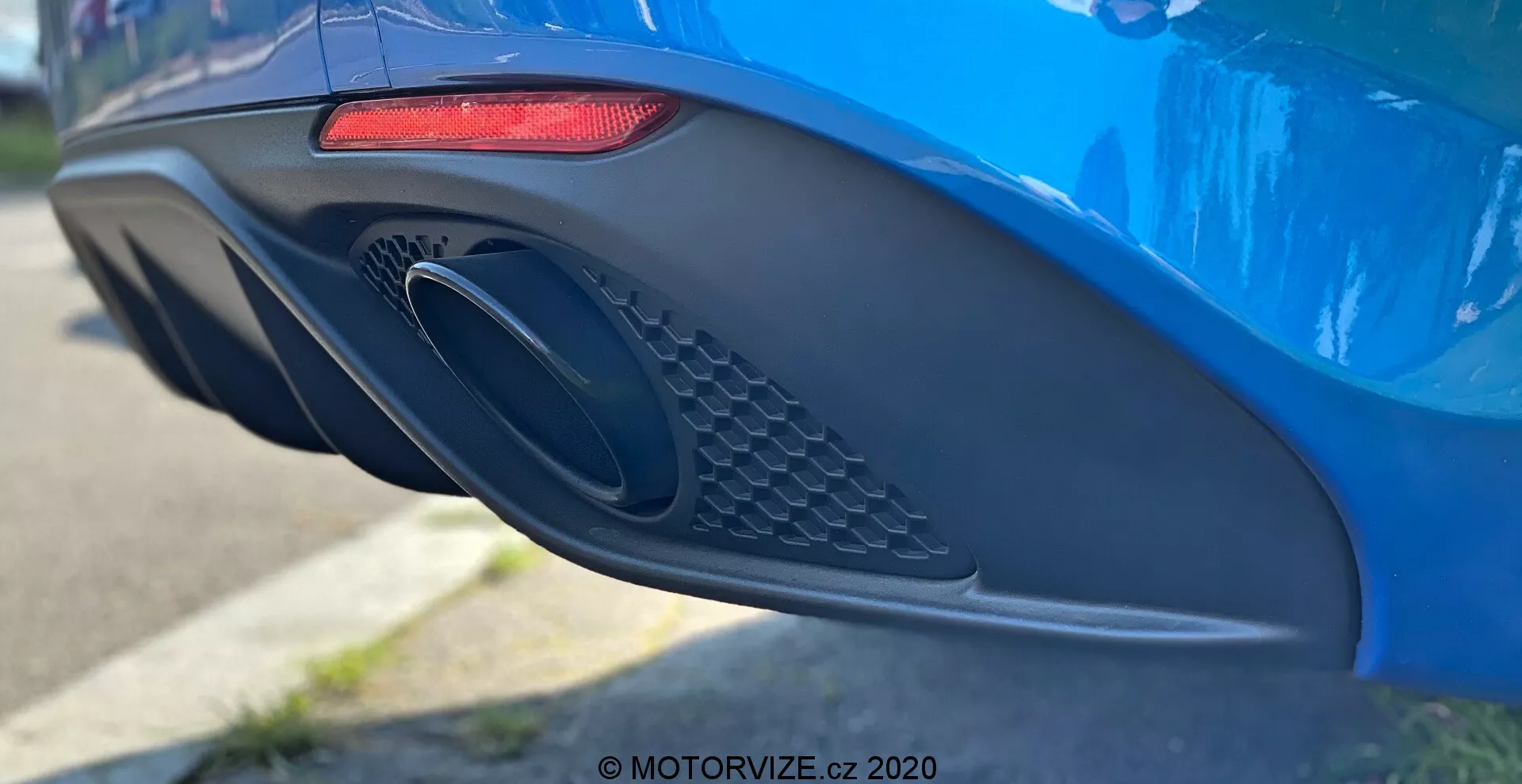 Vista lateral aproximada da área do para-choque traseiro e do escape de um vibrante Alfa Romeo Giulia 2016 azul, destacando suas características de design moderno, incluindo uma luz traseira LED horizontal vermelha alongada, uma grande porta de escape oval com detalhe de grade e um para-choques aerodinâmico com varias curvas e reentrâncias, fotografado com pequena profundidade de campo para enfatizar os detalhes do carro.