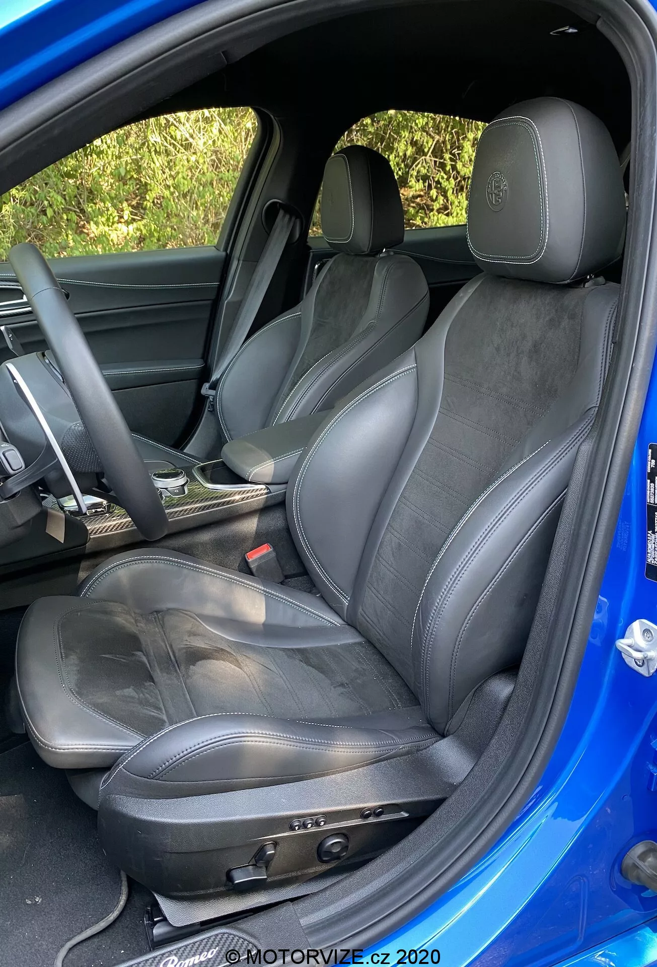 Vista detalhada do assento do motorista esportivo em um Alfa-Romeo Giulia 2016, mostrando ajustes manuais do assento, estofamento de couro e camurça, logotipo em relevo no apoio de cabeça, leve desgaste, volante parcialmente visível coberto de couro e acabamento de porta correspondente, capturado a partir de um ângulo externo acima do nível do assento com interior escuro e exterior azul brilhante contrastante.