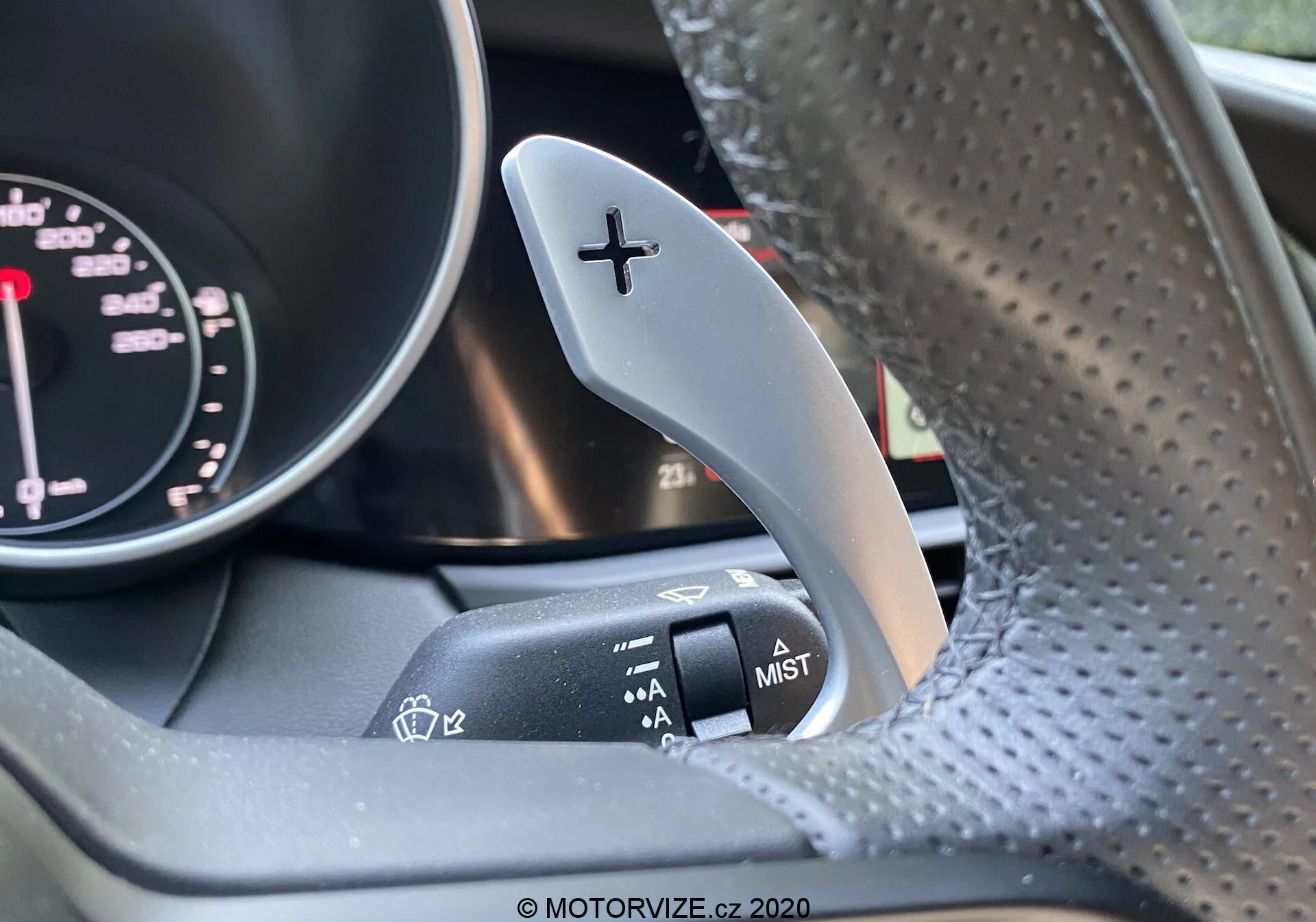 Close-up do interior de um Alfa Romeo Giulia 2016 focando na palheta de mudanças à direita com um sinal '+' para subir as marchas e a alavanca de controle do limpador de para-brisa adjacente na coluna de direção. Parte do tacômetro do painel de instrumentos está levemente visível. O volante, feito de couro perfurado, está um pouco desfocado. A imagem destaca o contraste entre a palheta de mudanças metálica e a alavanca de controle do limpador com acabamento fosco.