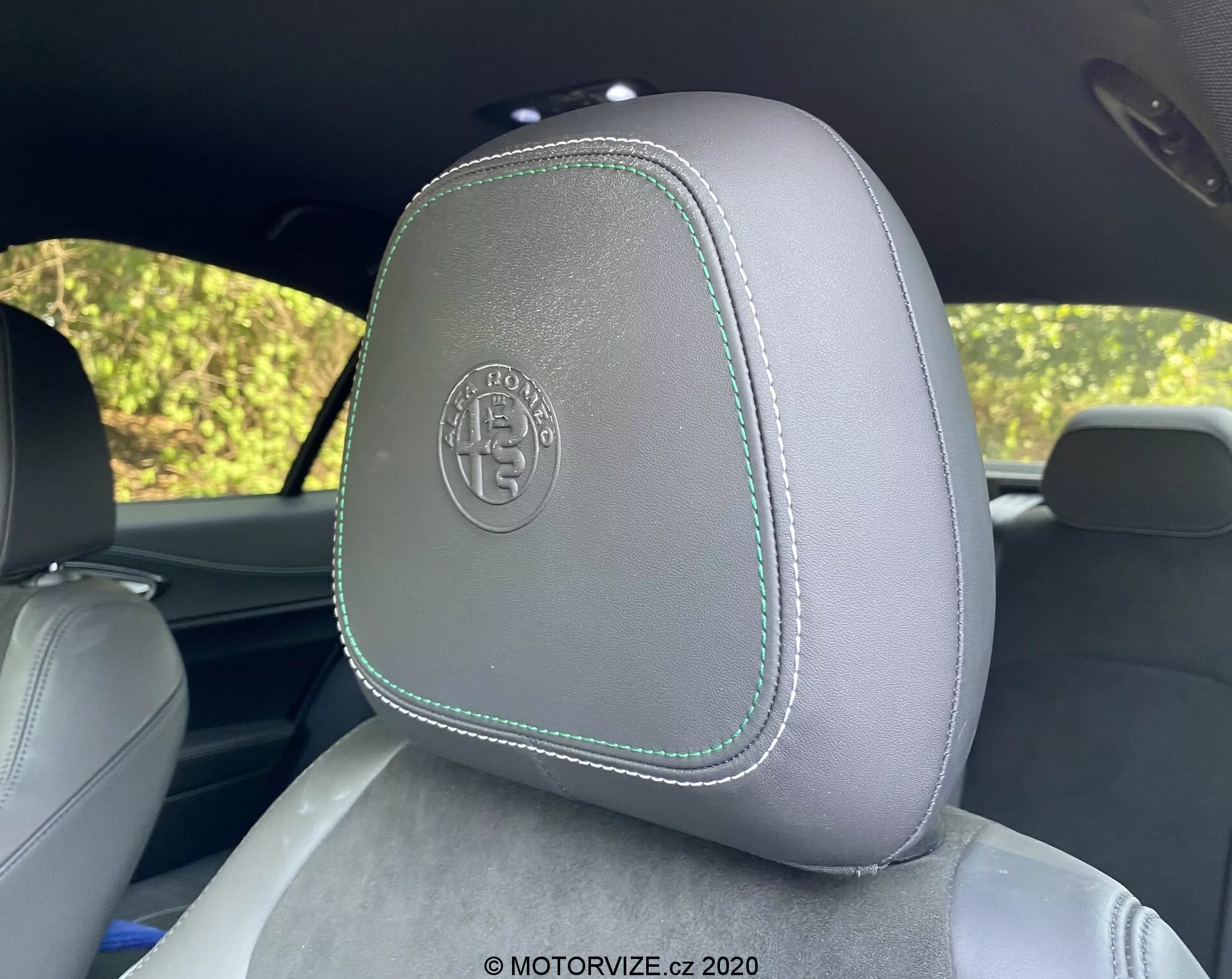 Close-up de um encosto de cabeça no Alfa Romeo Giulia 2016, mostrando estofamento de couro cinza claro com costuras contrastantes de cor verde claro, visto do banco traseiro. O encosto de cabeça apresenta um logotipo circular em relevo, sugerindo um acabamento premium ou personalizado. O fundo mostra folhagem desfocada através das janelas do carro, sugerindo um ambiente externo e claro.