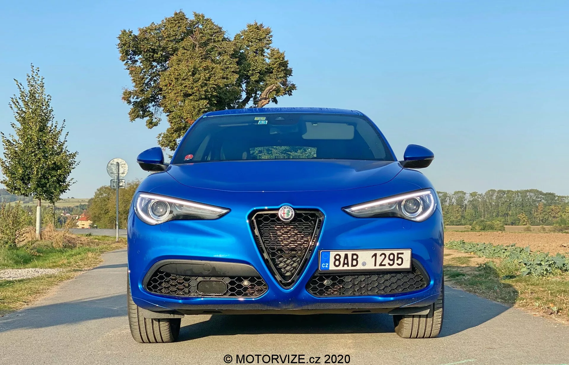 Vista frontal de um Alfa-Romeo Stelvio 2017 azul metálico, destacando sua postura agressiva e esportiva. As principais características incluem uma grade triangular grande e distintiva com o logotipo da marca, faróis elegantes com luzes diurnas, grandes entradas de ar no para-choque dianteiro e um capô com linhas elevadas. A pintura azul metálica indica um acabamento premium.