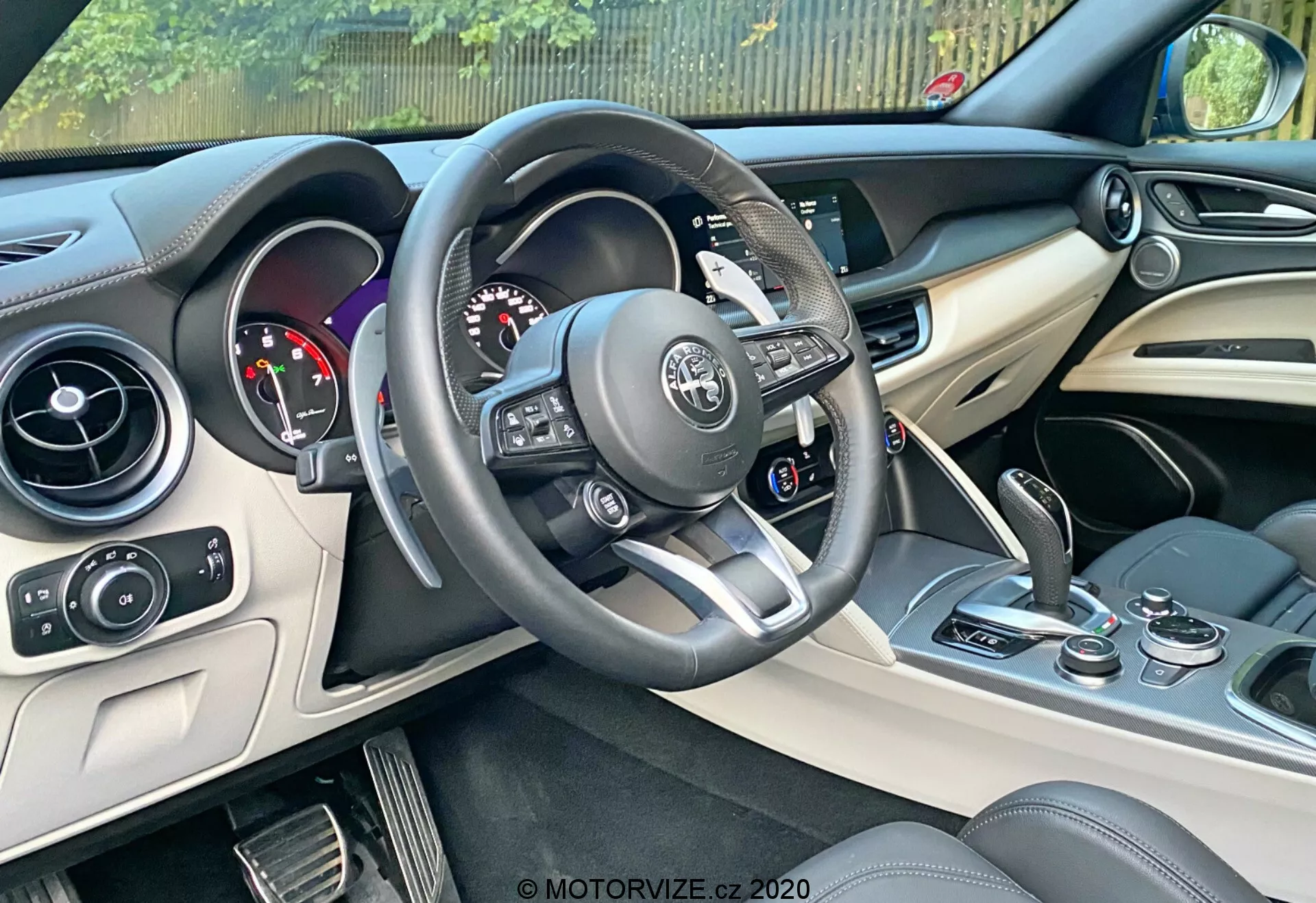 Foto do interior de um Alfa Romeo Stelvio 2017 focado no lado do motorista. Apresenta volante multifuncional revestido em couro com detalhes em prata e controles embutidos, painel de instrumentos com mostradores analógicos e visor digital, console central com sistema de infoentretenimento e controles de climatização, e uma visão dos bancos de couro em dois tons. Elementos de design moderno incluem detalhes em prata ou cromo no console e materiais luxuosos no painel e portas. Inclui botão de start/stop próximo ao console central.