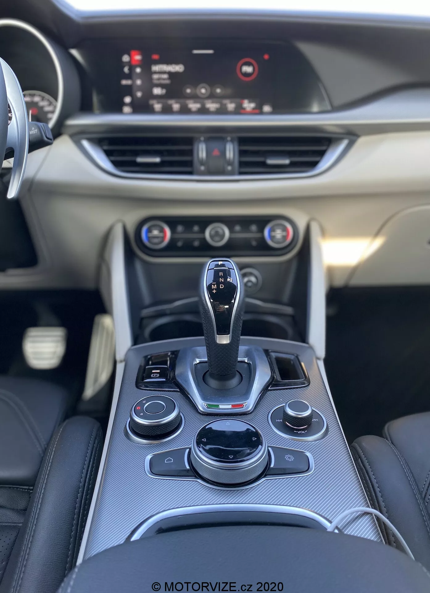 Vista interior do Alfa Romeo Stelvio 2017, mostrando o console central com uma alavanca de câmbio automática e uma disposição ergonômica de botões e dials, incluindo o sistema de infotainment, controles de climatização e possivelmente modos de condução. O painel inclui controles adicionais, saídas de ar e uma tela de infotainment destacada. Painel de passageiros liso com acabamento decorativo. Fotografia de alta qualidade destacando os materiais premium e o design elegante. © MOTORVIZE.cz