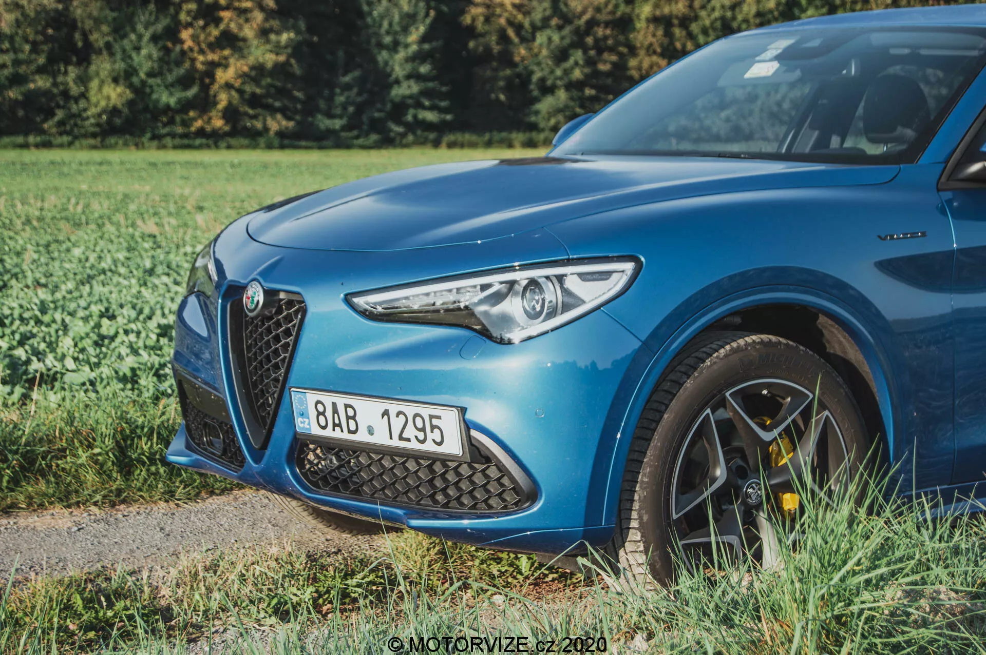 Um Alfa Romeo Stelvio azul de 2017 visto de um ângulo baixo de três quartos frontais, destacando o lado dianteiro esquerdo, incluindo o farol de LED moderno, a grande grade triangular intricada, o capô aerodinamicamente moldado e parte da roda de liga de múltiplos raios com pinças de freio de alto desempenho amarelas. O carro está parado sobre uma margem gramada ao lado de uma superfície pavimentada, com uma profundidade de campo rasa que desfoca o fundo de vegetação, enfatizando os detalhes nítidos do design e as características esportivas do carro.
