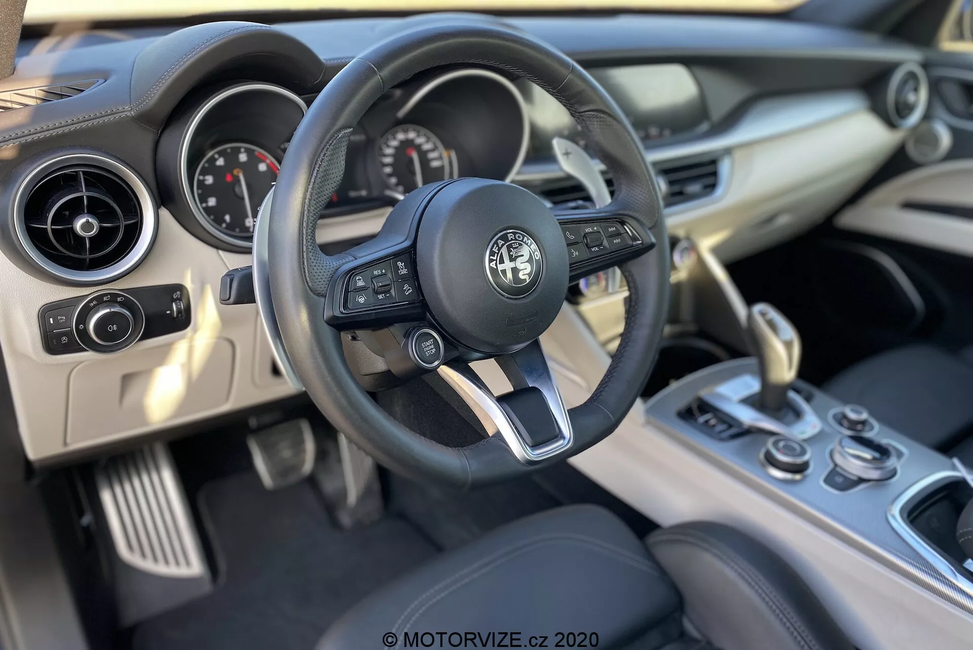 Vista interior do Alfa-Romeo Stelvio 2017 da perspectiva do motorista, destacando o volante com o emblema Alfa Romeo, controles de áudio e cruzeiro integrados, borboletas de mudança, tacômetro e velocímetro analógicos, display digital, ventiladores de ar circulares, painel de toque suave, botões do console central, botão rotativo e seletor de marchas de couro.