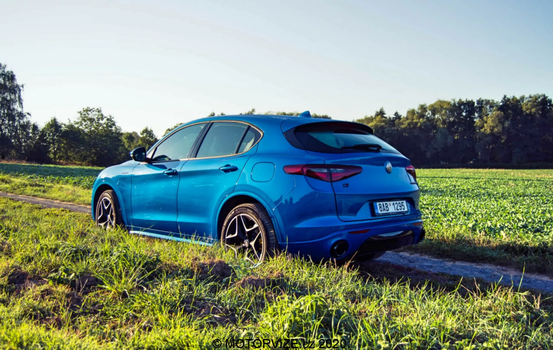 Imagem de um Alfa Romeo Stelvio azul de 2017, um crossover SUV esportivo moderno, tirada de uma perspectiva traseira de três quartos em um caminho de terra adjacente a um campo durante a hora dourada. Características incluem um spoiler esportivo, luzes traseiras elegantes, difusor traseiro com ponteiras de escape duplas, placa de registro central e emblema. A vista lateral mostra arcos de roda pronunciados, linha característica nas portas, grandes rodas de liga leve multirraio com pneus de perfil baixo, espelhos laterais da cor da carroceria e uma antena traseira. O Stelvio tem vidros traseiros escurecidos, uma linha de ombro forte e um efeito de lens flare do sol, destacando seu design dinâmico.