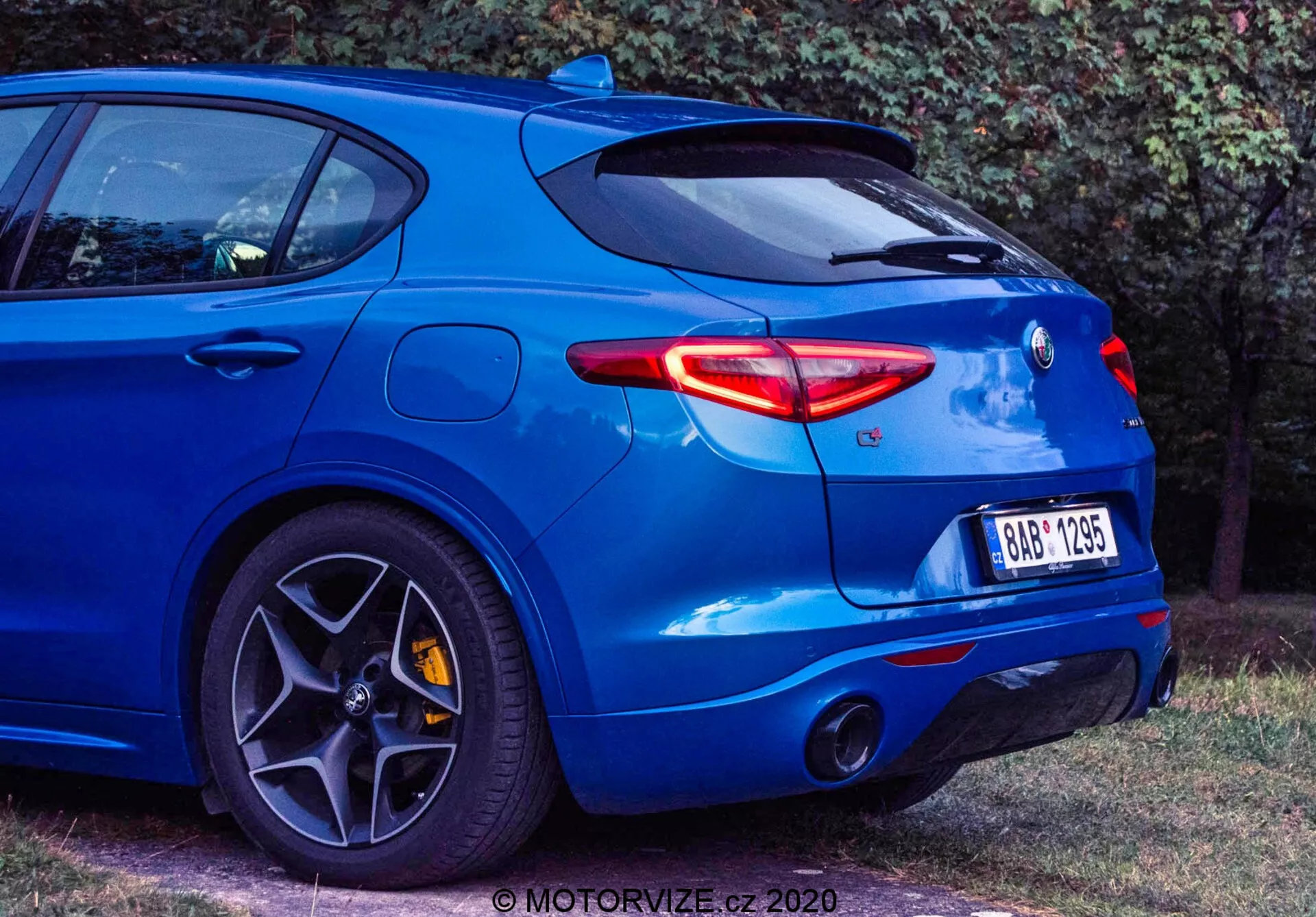 Vista traseira de três quartos de um hatchback azul Alfa Romeo Stelvio 2017, capturada de um ângulo baixo para mostrar suas características de design moderno, incluindo luzes traseiras LED elegantes, sistema de escape duplo, emblema traseiro centralizado, spoiler traseiro montado no teto, grandes rodas de liga leve multirraios com freios de alto desempenho, pneus de perfil baixo e carroçaria musculosa com para-lama traseiro saliente. O veículo exala uma estética esportiva e orientada para alta performance. Uma marca d'água de crédito é visível no canto inferior direito.