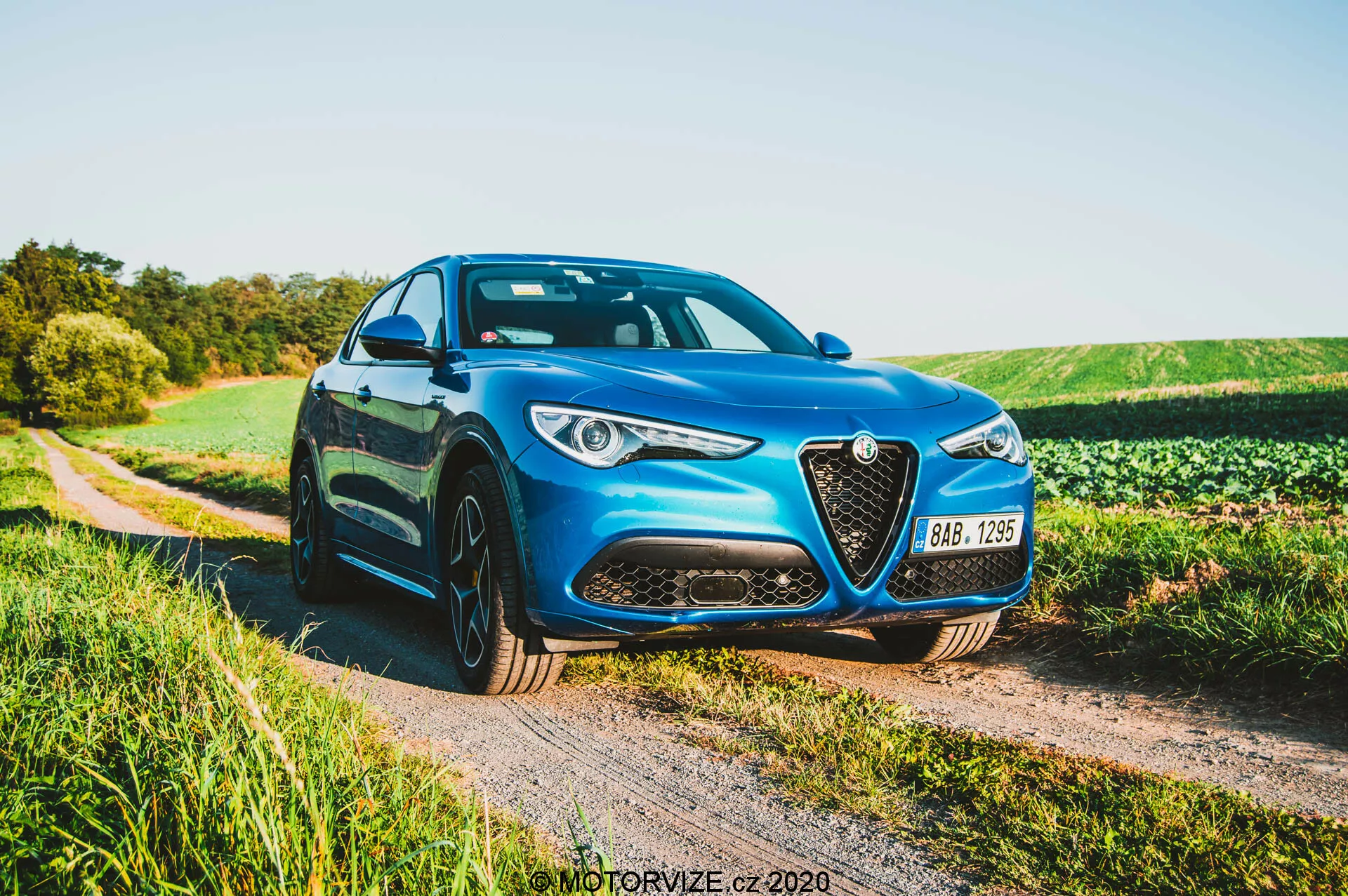 Alfa Romeo Stelvio 2017 azul estacionado em um caminho de cascalho ao lado de um campo no final da tarde, mostrando a grade frontal com contorno cromado, faróis de LED, linhas esculpidas no capô e rodas de liga leve multirraios em um ângulo dianteiro de três quartos.