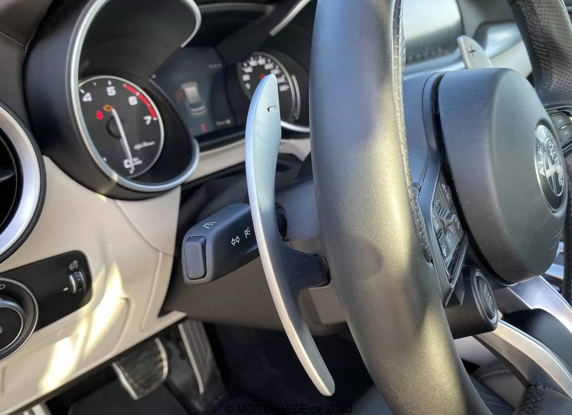 Vista interior de um Alfa-Romeo Stelvio 2017 a partir do assento do motorista, destacando o volante revestido de couro com botões multifuncionais, painel de instrumentos parcialmente visível com mostradores analógicos e um display digital central, e alavanca de controle à esquerda para as luzes exteriores. O painel apresenta aberturas de ar centrais com acentos cromados. Imagem bem iluminada mostrando a interface e os controles do motorista.