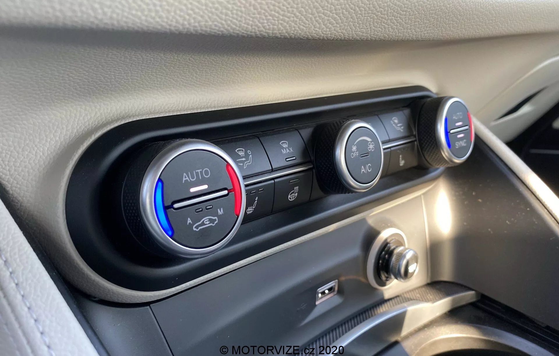 Visão aproximada do sistema de controle de climatização interior do Alfa Romeo Stelvio 2017. A imagem mostra os controles de temperatura e ventilador de um ângulo ligeiramente elevado, focando nos detalhes dos botões e controles. O controle de temperatura, ajustado em frio, possui uma faixa azul e vermelha, marcado com 'AUTO'. Ao lado, vários botões horizontais controlam a direção do fluxo de ar, recirculação de ar e desembaçamento do para-brisas. O controle à direita indica a velocidade do ventilador com luzes LED, ajustado para baixa. Abaixo, destaca-se um botão 'A/C'. O painel de controle está emoldurado em um entorno preto brilhante, contrastando com o painel claro, capturado em alta resolução com reflexos claros de luz natural.