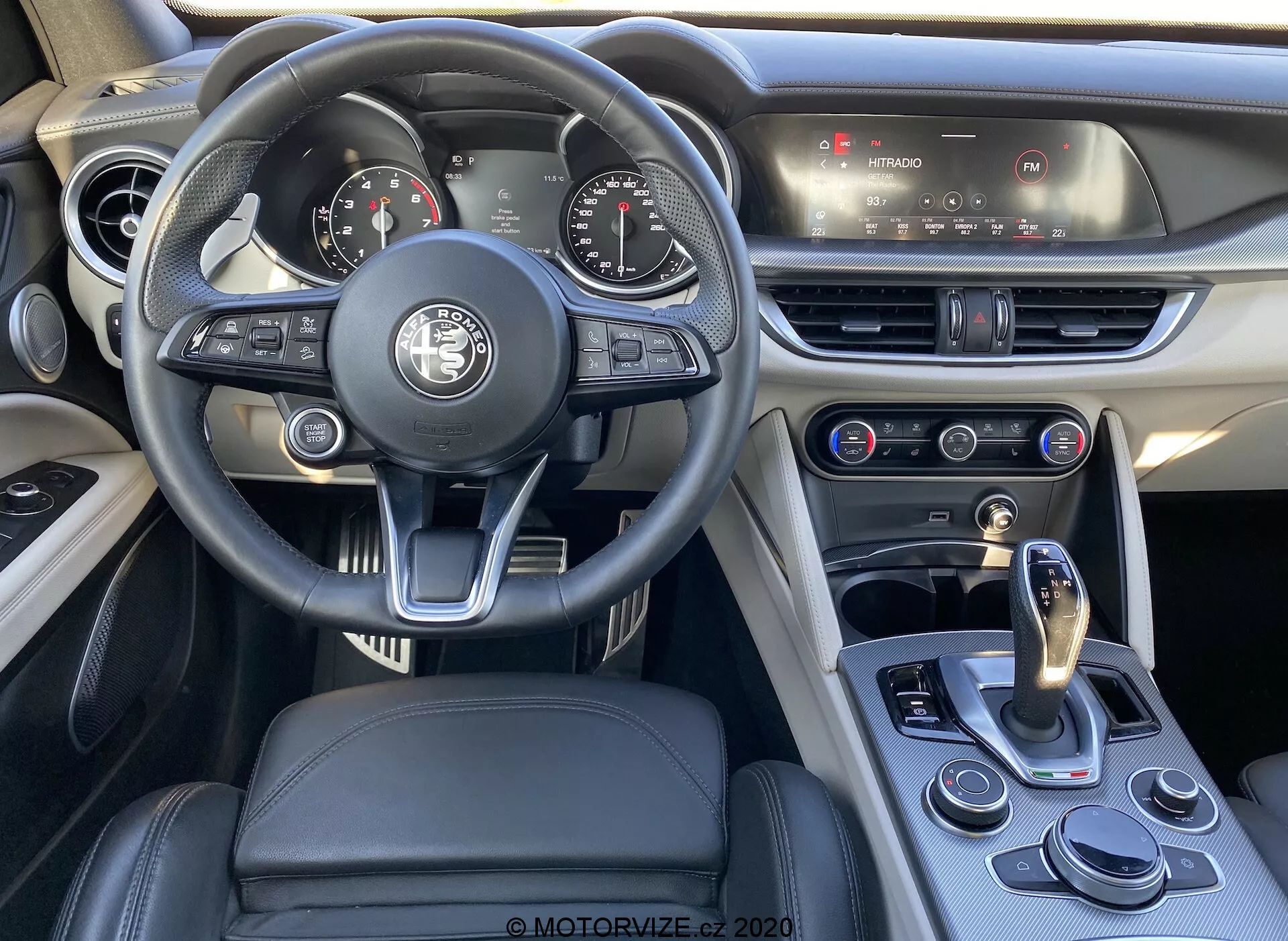 Uma imagem do interior de um Alfa Romeo Stelvio 2017, focando na área do motorista, destacando o volante multifuncional moderno revestido em couro com vários botões de controle, um painel de instrumentos analógico-digital parcialmente visível e o console central com sistema de infotainment touchscreen e controle de clima. O design combina luxo e esportividade com estofamento de couro e costuras contrastantes. A alavanca de câmbio e o seletor de modos de condução destacam a transmissão automática e os modos de condução selecionáveis do veículo.