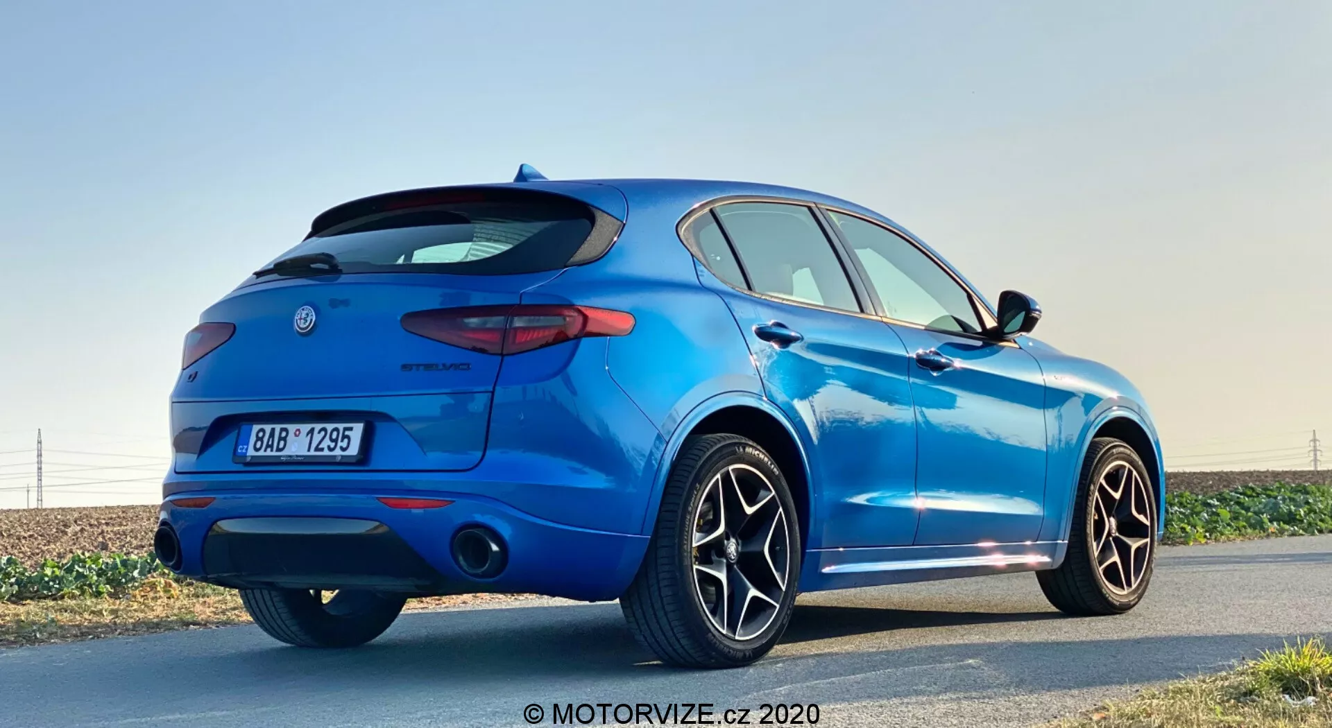 Vista traseira de três quartos de um SUV compacto azul Alfa Romeo Stelvio 2017 estacionado ao lado de uma estrada à luz do dia com céu claro, destacando o spoiler traseiro esportivo, luzes traseiras de LED, sistema de escape duplo, grandes rodas de liga leve com múltiplos raios, sensores de estacionamento e antena tipo barbatana de tubarão, em um cenário que realça seu design elegante e características modernas.