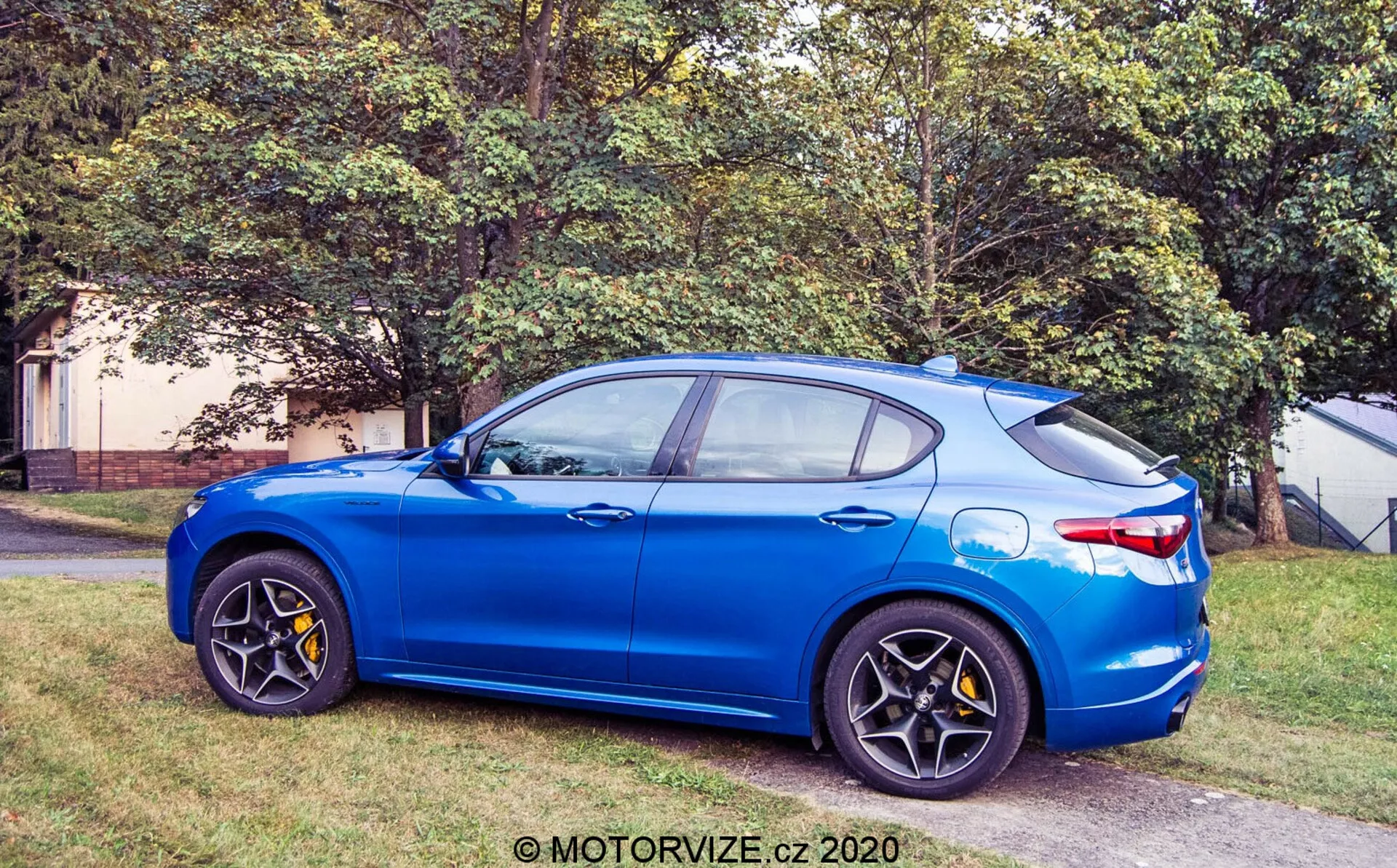 Vista de perfil lateral do Alfa Romeo Stelvio 2017 em azul brilhante estacionado em uma área gramada ao lado de uma superfície pavimentada, mostrando a visão do quarto dianteiro com rodas de liga leve preto de múltiplos raios, pinças de freio amarelas, espelhos laterais na cor da carroceria, linha do teto inclinada com antena de barbatana de tubarão e design de quatro portas com acabamento metálico.