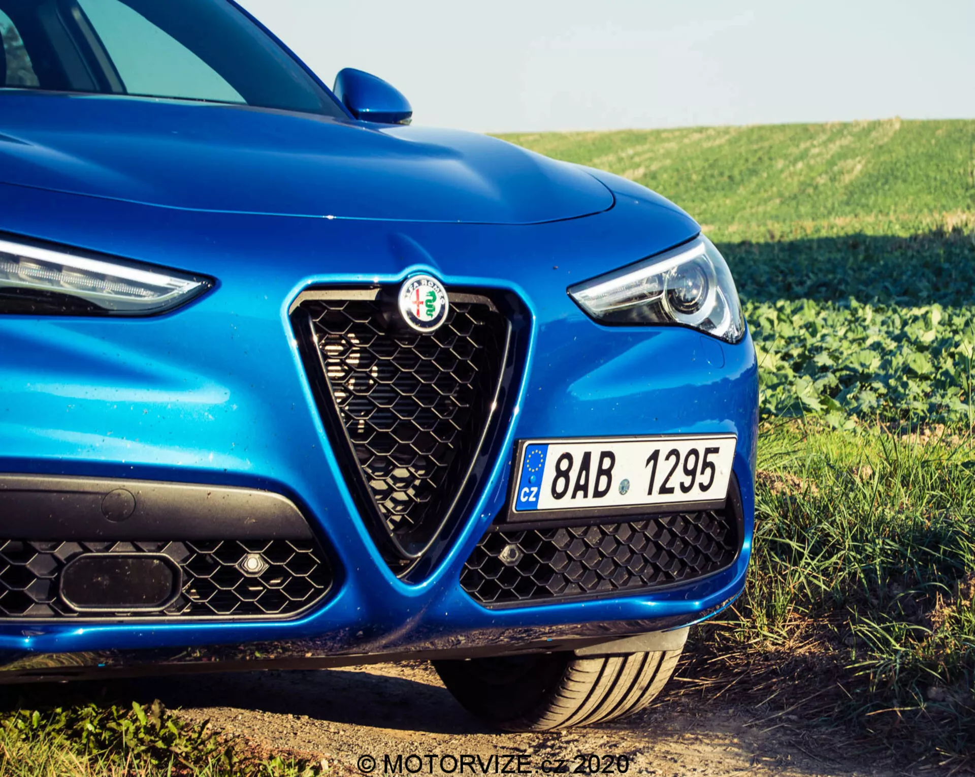 Close-up do lado frontal esquerdo de um Alfa Romeo Stelvio 2017 azul, mostrando o emblema distintivo na grade hexagonal, o design elegante do farol com elementos LED e parte do capô. A imagem, tirada de um ângulo baixo, destaca a frente esportiva e agressiva do veículo, incluindo o para-choque dianteiro preto com grandes entradas de ar e uma placa de licença checa.