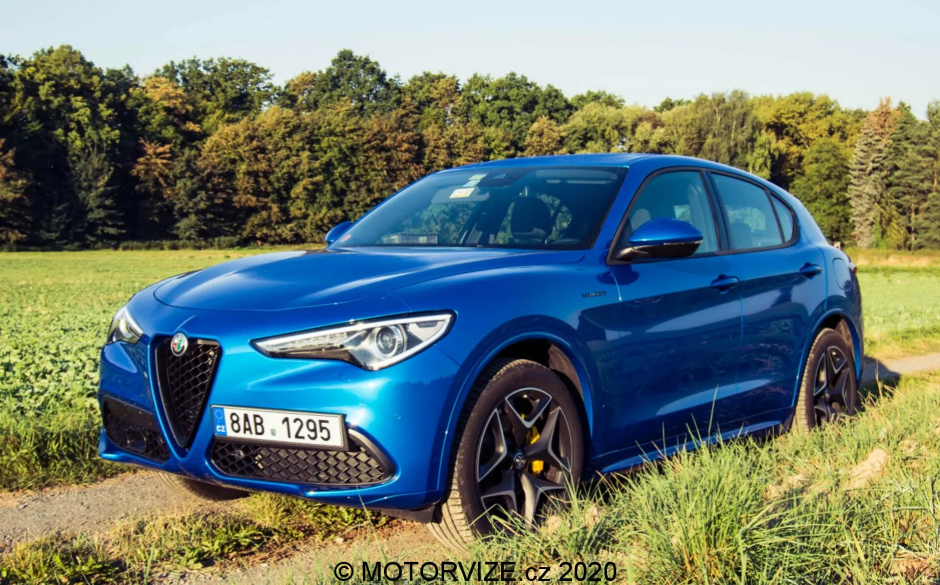 Vista em ângulo três-quartos frontal do Alfa Romeo Stelvio 2017 em metálico azul vibrante, destacando a grade triangular “Scudetto” característica, os faróis LED elegantes, o design aerodinâmico do capô com linhas de vinco distintas, as rodas de liga leve de vários raios de cor escura, os pneus de perfil baixo de alto desempenho, os espelhos laterais na cor da carroceria com setas integradas e as molduras das janelas na cor da carroceria com o pilar B preto. O veículo está estacionado em um caminho não pavimentado ao lado de um campo de grama sob um céu claro com sombras suaves, sugerindo luz de manhã cedo ou final de tarde.