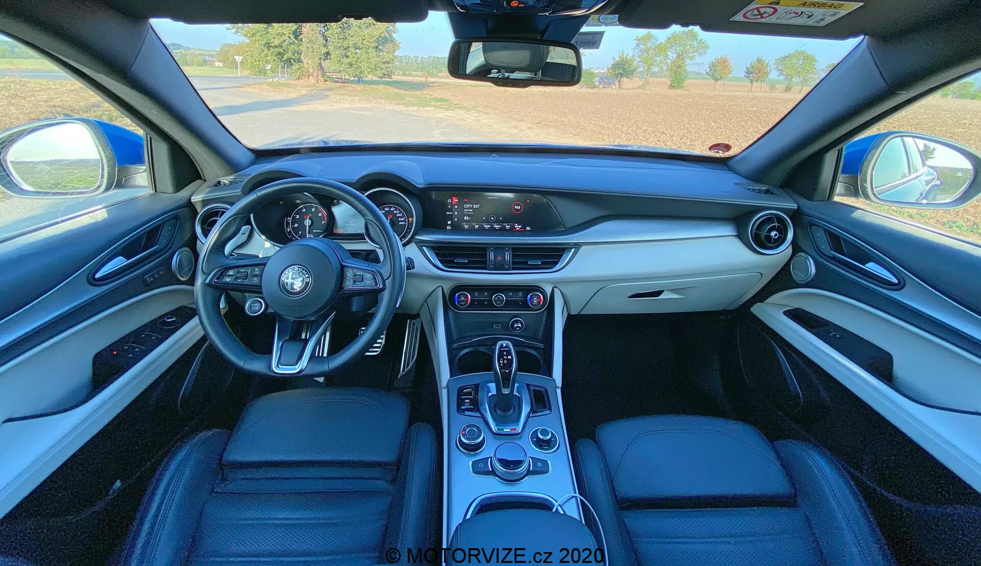 Vista interior de um Alfa Romeo Stelvio 2017 a partir de uma perspectiva em primeira pessoa, mostrando o painel, volante de três raios com controles montados, centro de informações digital do motorista, sistema de infotainment com tela sensível ao toque, console central com alavanca de câmbio automática e estofamento de couro premium com materiais contrastantes.