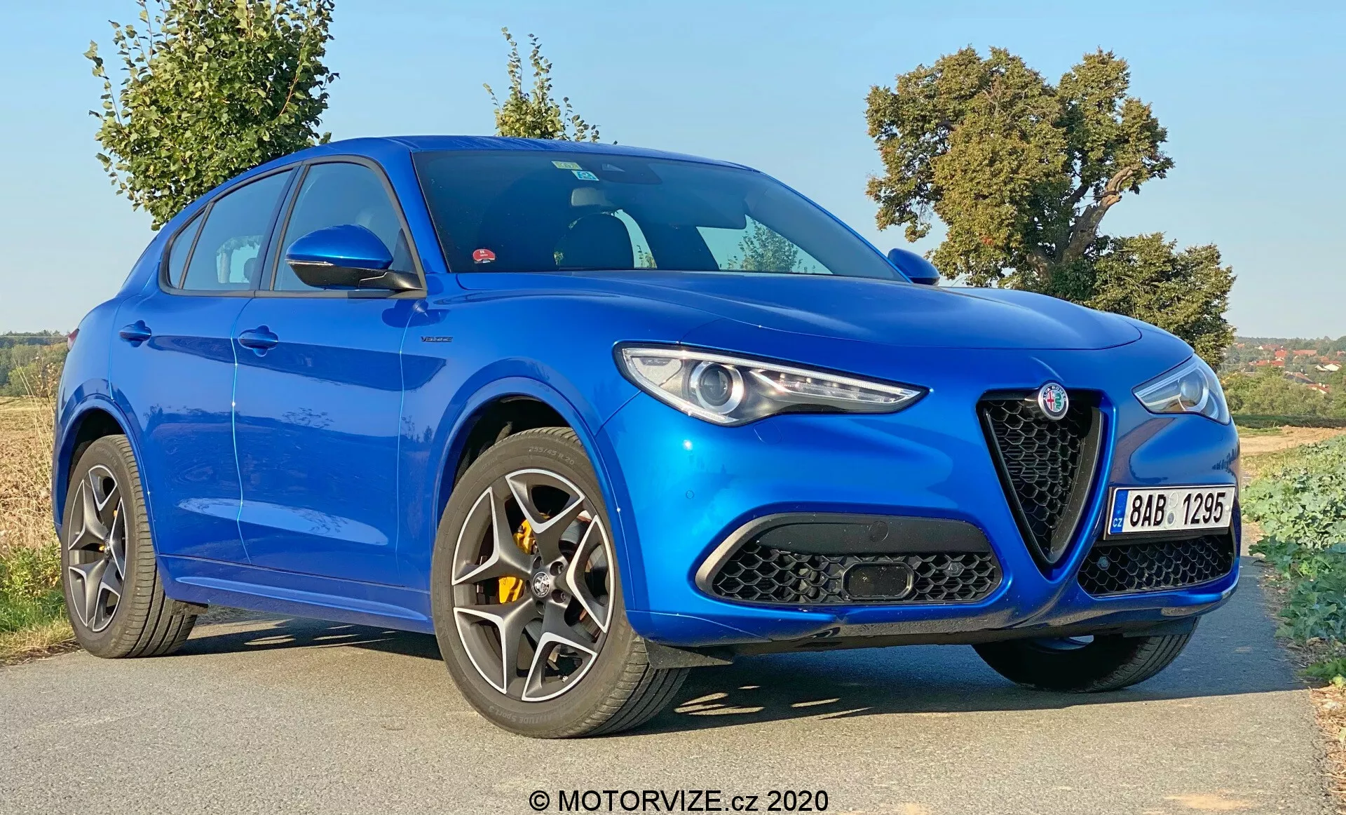 Vista frontal de três quartos do Alfa Romeo Stelvio 2017 em azul brilhante, destacando sua distinta grade triangular com um logotipo proeminente, faróis estreitos e elegantes, contornos pronunciados do capô e rodas de múltiplos raios. Esta imagem capta o design agressivo e moderno do veículo com grandes entradas de ar no para-choques dianteiro, uma silhueta esportiva com uma linha de teto descendente e linhas esculpidas no corpo, sugerindo uma identidade orientada para desempenho e luxo.