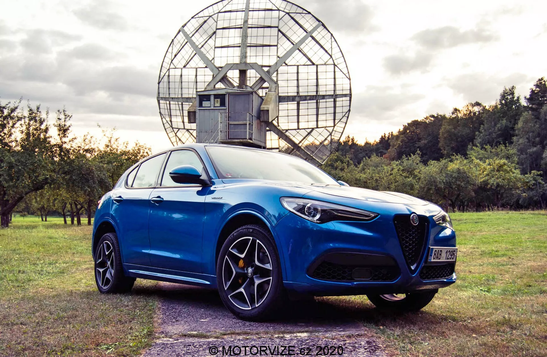 Foto de um sedan esportivo moderno azul capturada a partir de um ângulo de três quartos da frente baixo, mostrando o perfil frontal e lateral. O carro apresenta uma grade triangular distinta, faróis angulares e afiados, e rodas de liga leve de múltiplos raios com pinças de freio visíveis. Está estacionado em uma área gramada com árvores e uma estrutura esférica de aparência industrial ao fundo. A foto é composta sob luz natural, destacando as linhas aerodinâmicas e o design suave do carro.
