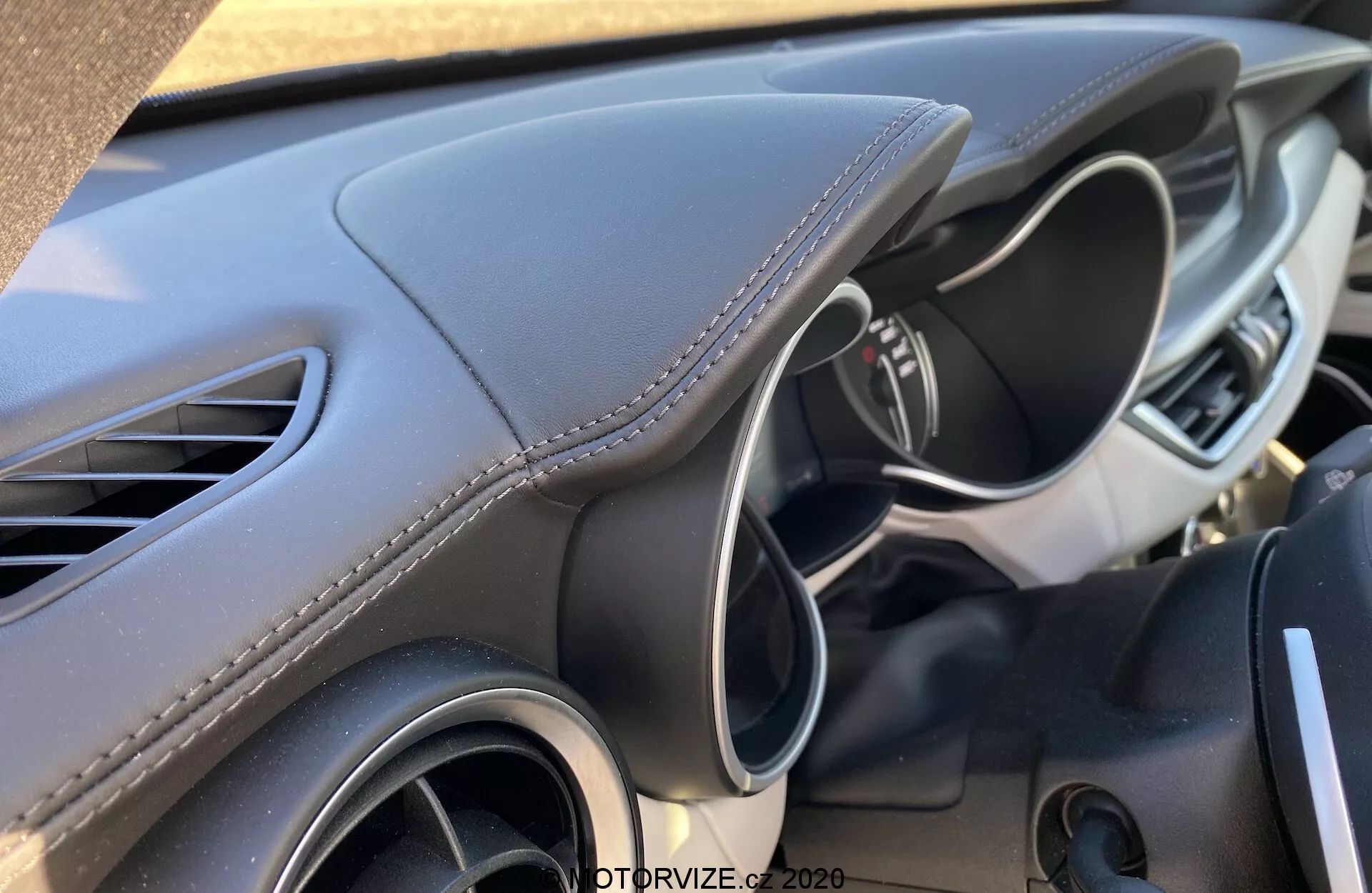 Vista de perto do interior do Alfa-Romeo Stelvio 2017, focando no lado do motorista do painel, visível do banco do passageiro ou da consola central. O design moderno e esculpido do painel apresenta material macio ao toque com costura branca, sugerindo uma estética premium. A saída de ar esquerda e parte do volante multifuncional com controles ou botões também são visíveis, sugerindo um ambiente avançado e amigável para o motorista.