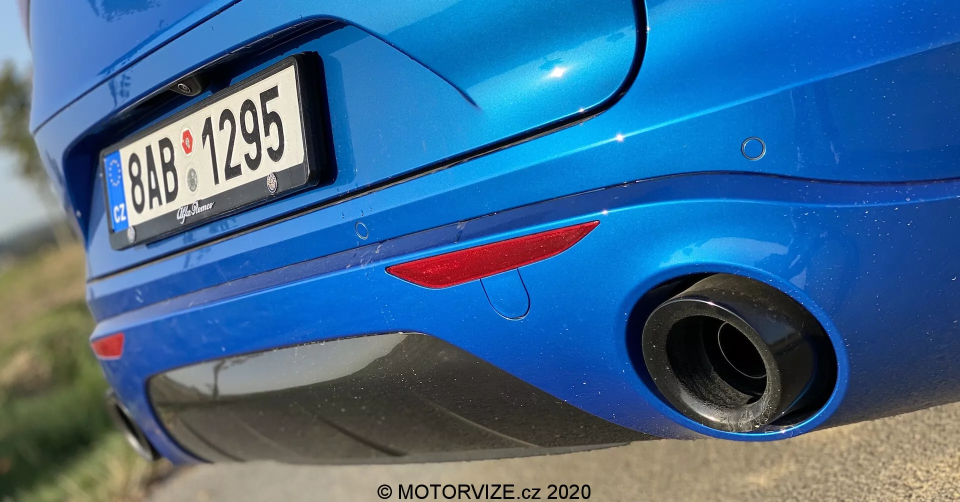 Vista aproximada da seção traseira de um Alfa Romeo Stelvio 2017 azul, tirada de um ângulo baixo. O tiro destaca a metade inferior da parte traseira do carro, focando no tubo de escape e no para-choques traseiro. As características incluem uma luz refletora traseira vermelha, um pneu traseiro parcialmente visível, um para-choques traseiro arredondado e brilhante, uma placa de licença parcialmente cortada, um tubo de escape arredondado com ponta cromada e uma vinco acima do escape no para-choques. A pintura metálica com reflexão da luz solar enfatiza a condição limpa e bem mantida do carro.