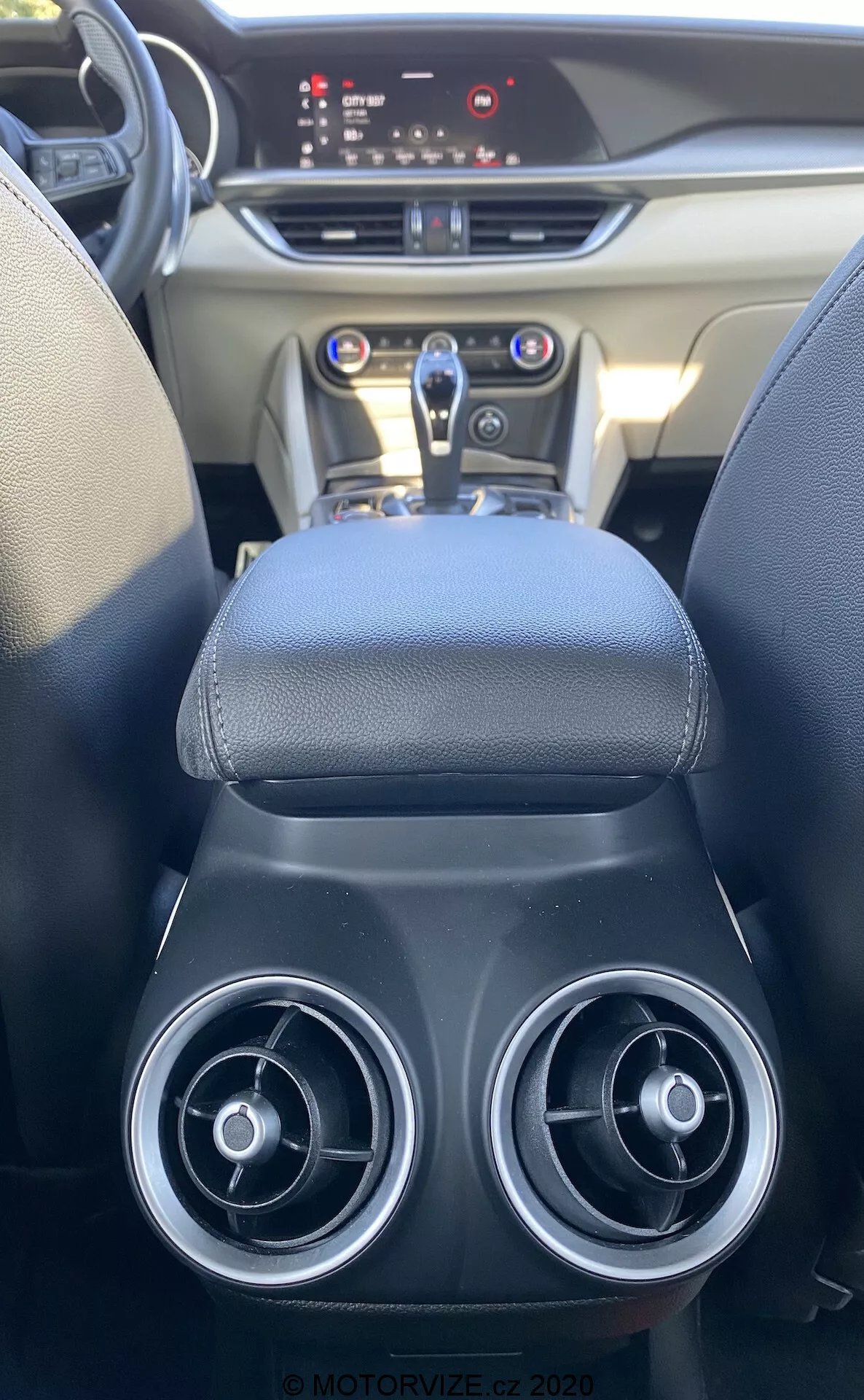 Vista interior do assento traseiro de um Alfa Romeo Stelvio 2017, focando na consola central e painel do carro. A imagem mostra duas saídas de ar circulares, um apoio de braço em couro, uma alavanca de câmbio automática, painel de controle brilhante com controles metálicos e um painel digital iluminado, destacando design sofisticado e recursos modernos.