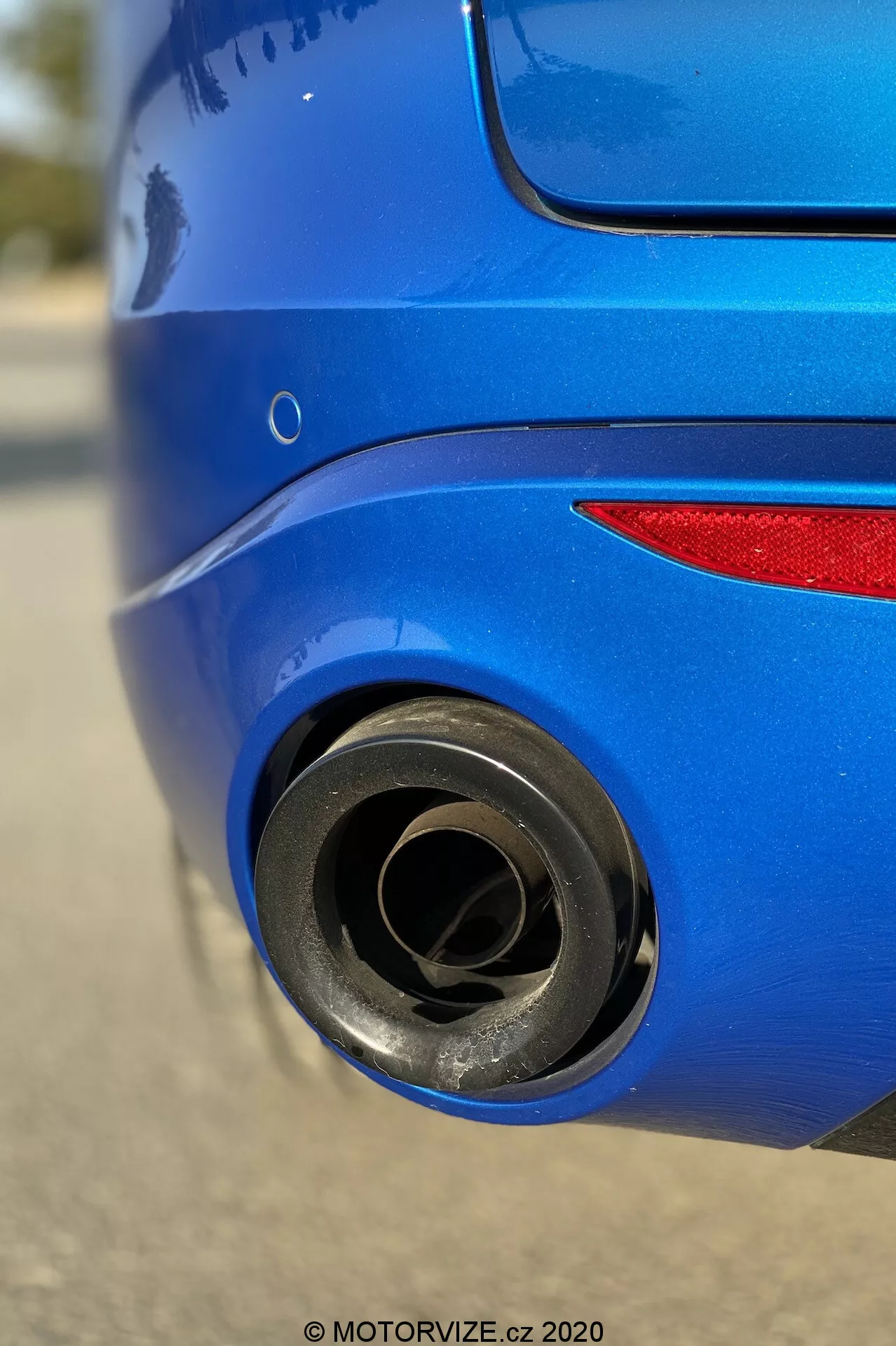 Close-up da seção traseira de um Alfa Romeo Stelvio 2017 azul, com foco no tubo de escape metálico com depósitos de fuligem visíveis, capturado de um ângulo baixo. A pintura azul refletora do para-choque traseiro e parte da luz traseira contrastam com a textura mate do tubo de escape, destacando as linhas de design e as características de desempenho do veículo.