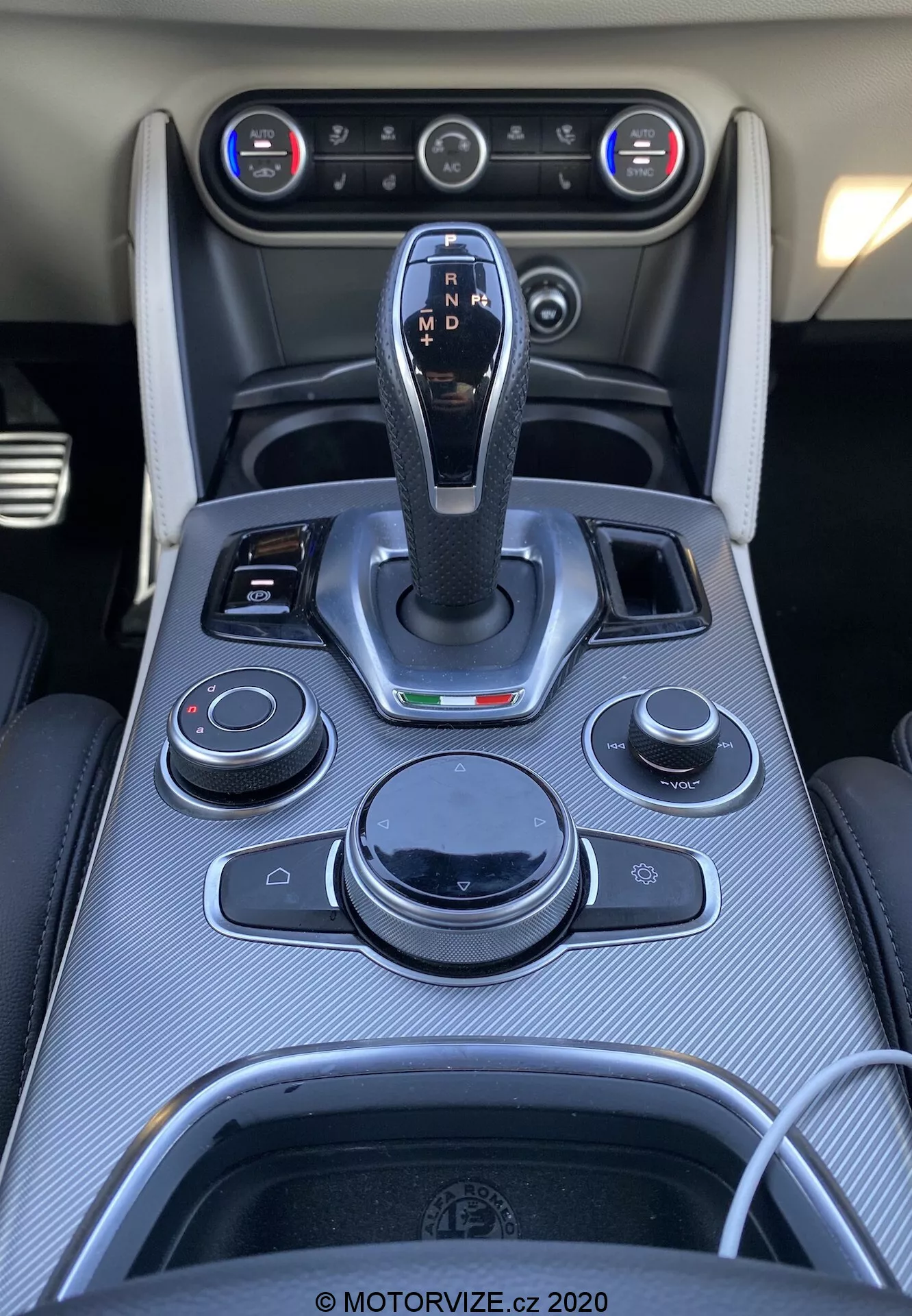 Vista de cima em close-up do console central no interior de um Alfa Romeo Stelvio 2017. Alavanca de câmbio central com acabamento em couro e detalhes cromados, identificada com configurações de transmissão automática. Botões de controle circundantes e interruptores para sistema de infoentretenimento, áudio, clima e funções do veículo. Acabamento em fibra de carbono, saídas de ar condicionado e sistema de controle de clima dual-zone visíveis. Porta-copos com contorno cromado e uma porta de conectividade presente.