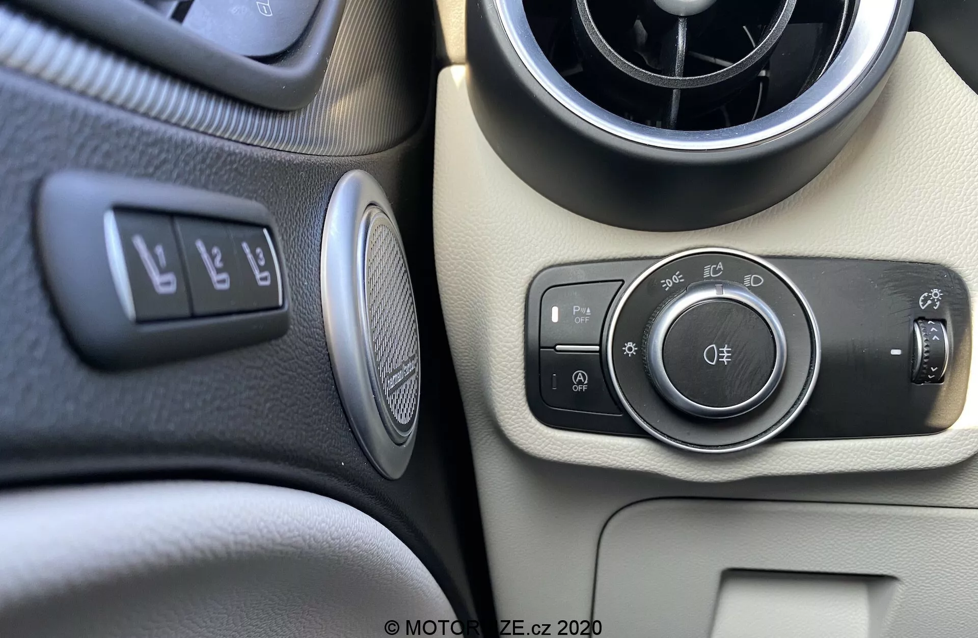Close-up do módulo de controle de iluminação e painel do Alfa Romeo Stelvio 2017 na perspectiva do motorista ou passageiro dianteiro. A imagem mostra os controles baseados em botões para faróis, luzes de estacionamento, iluminação automática e faróis de neblina dianteiros e traseiros. Ao lado, há uma alavanca para ajustar o ângulo dos faróis. Elementos visíveis incluem uma ventilação circular, um painel de dois tons em preto e bege, botões para controle de tração, abertura do porta-malas, assistência de estacionamento, um emblema circular provavelmente representando a marca do carro ou o sistema de áudio e uma grade de alto-falante arredondada.